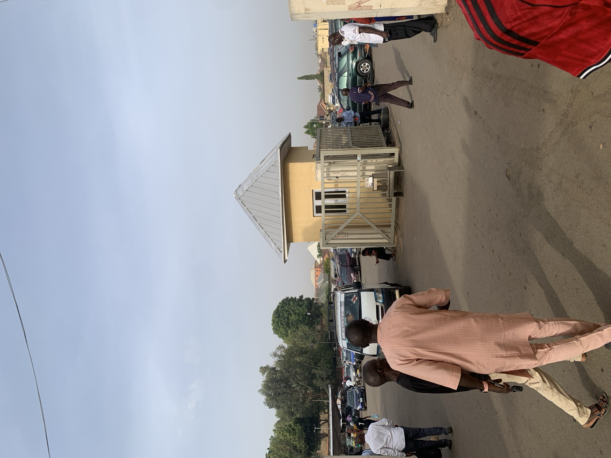 This image depicts a daytime street scene in Bukuru, Nigeria, filled with people and vehicles under a clear sky. The perspective is slightly elevated, looking down a paved road.
In the immediate foreground, a man wearing a light pink, striped traditional Nigerian outfit (a long tunic and matching trousers) and sandals stands with his back largely to the camera. Adjacent to him, another person is seen in a black top and khaki trousers. Throughout the scene, numerous individuals, predominantly adult males, are visible standing, walking, and congregating along the road and near the vehicles. One man in the midground wears a white and black striped short-sleeved top.
A prominent feature in the midground is a small, light-yellow building with a distinctive grey, pyramidal metallic roof. This structure appears to be a gatehouse or security post, featuring a white-framed window and a metal gate/fence to its right.
The road is occupied by several vehicles. A dark green SUV is parked near the gatehouse. Further down, a white van or bus is clearly visible, with the word "TOYOTA" prominently displayed on its side. Other cars are parked or moving in the background, and green trees line the street in the distance. The overall lighting suggests it is daytime, with visible shadows indicating either late morning or afternoon. The scene conveys the ordinary hustle and bustle of a local community street.