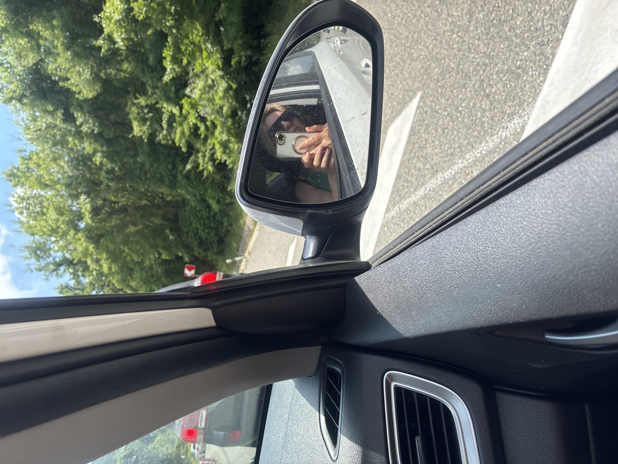 The image is taken from inside a car, looking out the passenger side window and into the side mirror. The side mirror reflects a woman wearing sunglasses and a green shirt, holding a white smartphone up to take a selfie. She is in the passenger seat of the car. Outside the car, the scenery consists of trees lining a road, and other cars are visible in the distance, suggesting traffic. A red car is partially visible in the foreground through the front windshield. The weather appears to be sunny with a blue sky and some clouds. The image is focused on the interior of the car and the reflection in the side mirror, with the exterior environment slightly out of focus. The location is identified as Arkhipo-Osipovka, Russia, a coastal resort town.