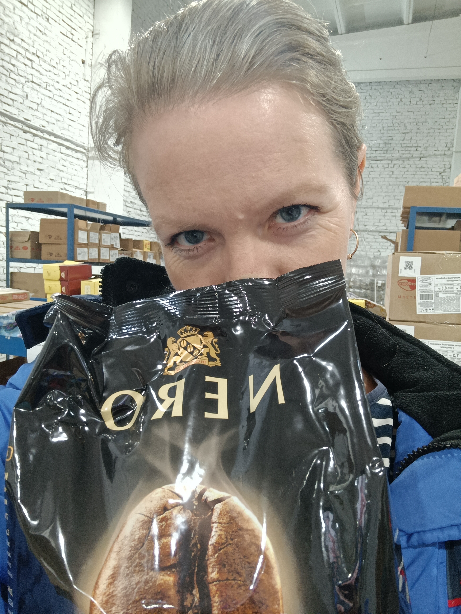 A woman with light, possibly graying hair and blue eyes is pictured indoors in Chaikovsky, Russia, taking a close-up shot. Her face is partially obscured by a large, glossy black bag she holds up, looking directly at the camera with an intense gaze and slightly furrowed brows. She is wearing a blue jacket over a striped shirt.
The black bag features gold lettering that reads "OЯƎN" (mirrored, indicating "NERO") and a golden emblem depicting a crown flanked by two lions. A large, detailed image of a roasted coffee bean is visible on the lower part of the bag.
The background suggests a storage or warehouse environment, with white painted brick walls and metal shelving units stacked with various cardboard boxes. Some smaller yellow and red packages are also visible on the shelves. The lighting appears to be artificial, typical of an indoor commercial setting, making the time of day undeterminable. No specific weather conditions can be inferred.