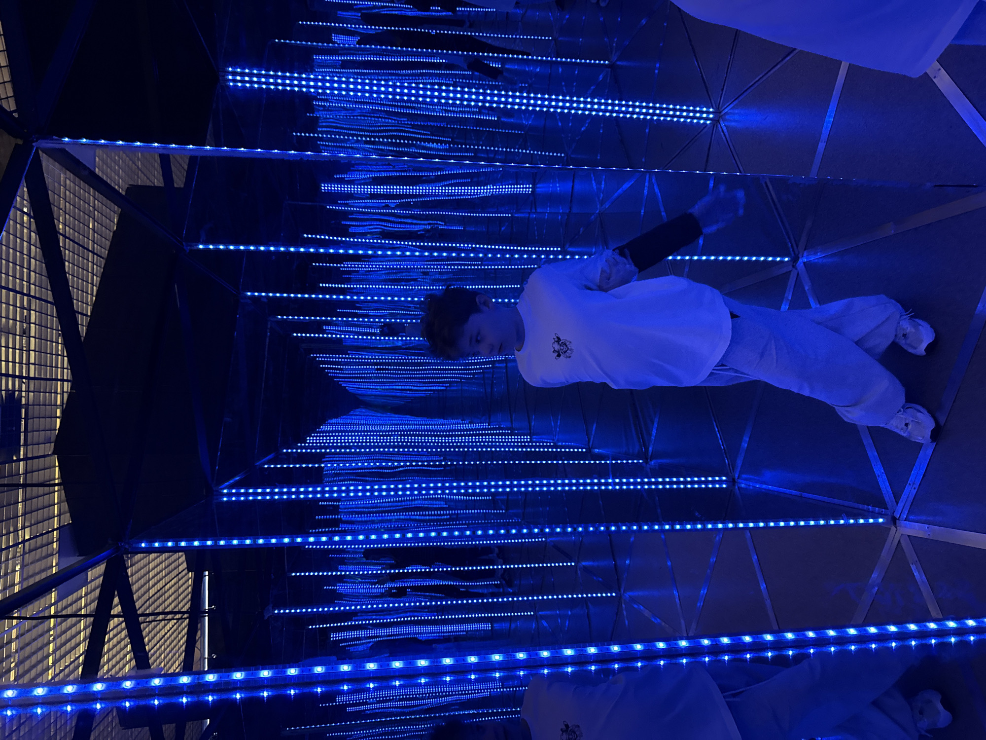 A young male is standing in a geodesic dome mirrored room featuring endless rows of blue LED strip lights. The framework of the dome is visible, creating triangular segments on the mirrored walls and ceilings. The male is wearing a white shirt, gray pants, and white sneakers. The location appears to be in Moscow, Russia.
