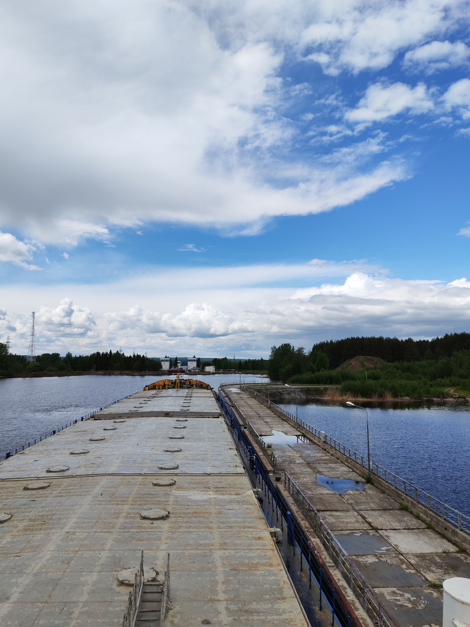 The image depicts a wide-angle view from the flat deck of a barge or ship, merging with a concrete pier or dock in Belousovo, Russia. The deck is grey, metallic, and has numerous circular access hatches. To the right of the deck, a concrete pier extends towards the horizon. The pier consists of rectangular slabs with metal railing. There are puddles of water on the pier. The scene is bordered by water, with forested areas on either side. The sky is a mix of blue and white clouds. In the distance, there appears to be a structure with tall white buildings. A transmission tower is visible on the left shore. The overall impression is of an industrial waterway setting on a cloudy day.