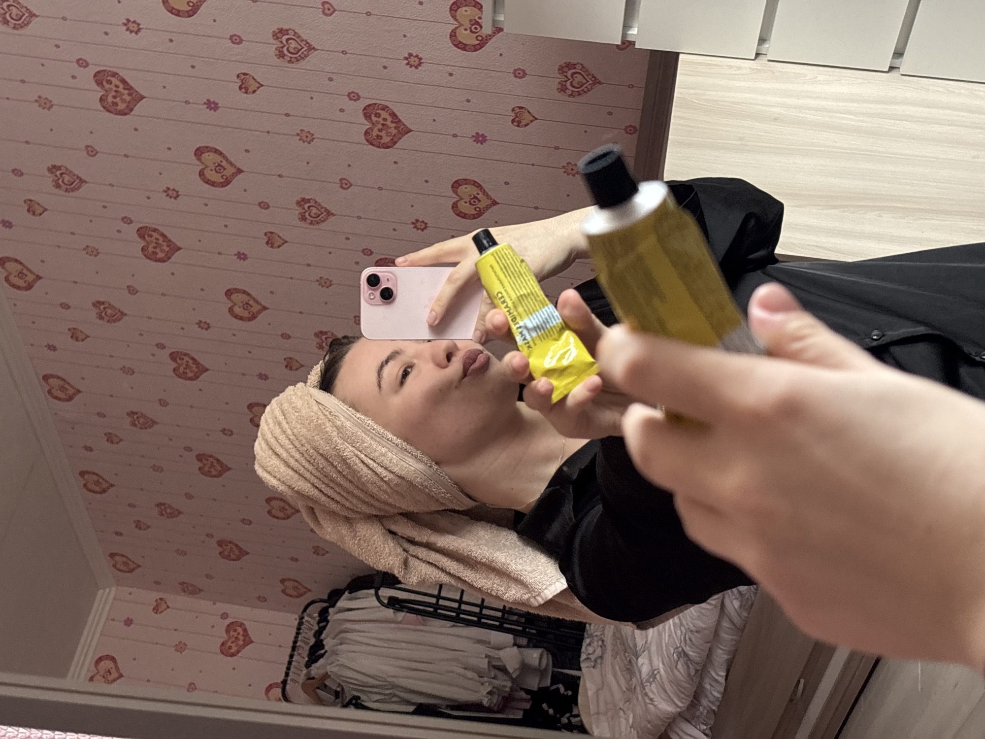 A woman is taking a selfie in a mirror. She is wearing a towel wrapped around her head and a black garment. She is holding a yellow tube of cream and a pink phone. The background includes a wall with a heart pattern, a clothes rack with clothes, and a light-colored wall. The woman is making a kissing face. The city of Perm, Russia is not discernible from the image.