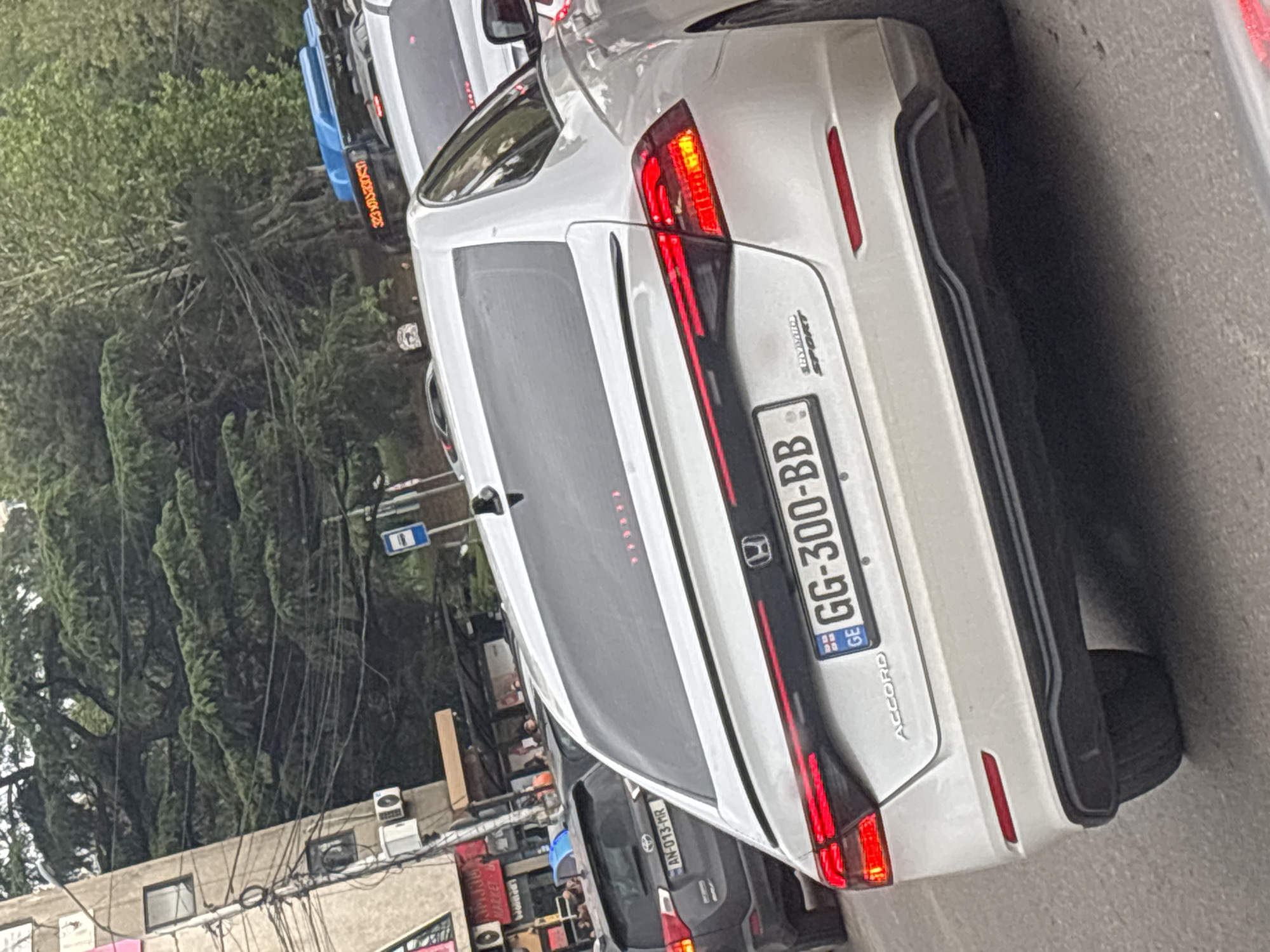 The image shows the rear view of a white Honda Accord Hybrid Sport car in Tbilisi, Georgia, with license plate GG-300-BB. The vehicle is positioned on a city street, presumably in traffic, as there are other vehicles visible in the background, including a grey car with license plate AN-013-MR. The photo shows a mix of urban and natural elements, with building and street infrastructure juxtaposed against dense trees and overgrown vegetation. There are also electrical wires running above the buildings.