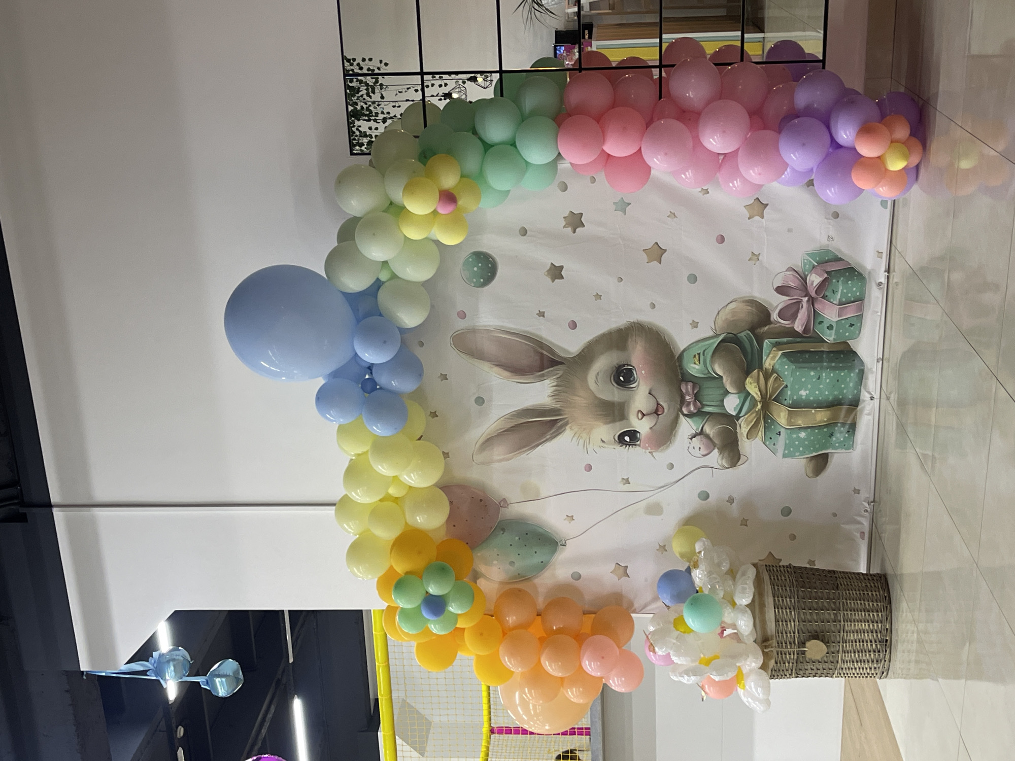 The image shows a colorful balloon arch with a bunny-themed backdrop. The arch is made of balloons in various pastel colors including blue, yellow, green, pink, and orange. A large, cartoonish bunny character is printed on the backdrop, holding an egg and standing next to two gift boxes. A woven basket filled with white floral-shaped balloons is placed to the left of the backdrop. The scene appears to be set up for a children's party or event. The location is identified as being in Donskoye, Russia. The background includes elements of an indoor space with white walls and a ceiling with lighting fixtures.