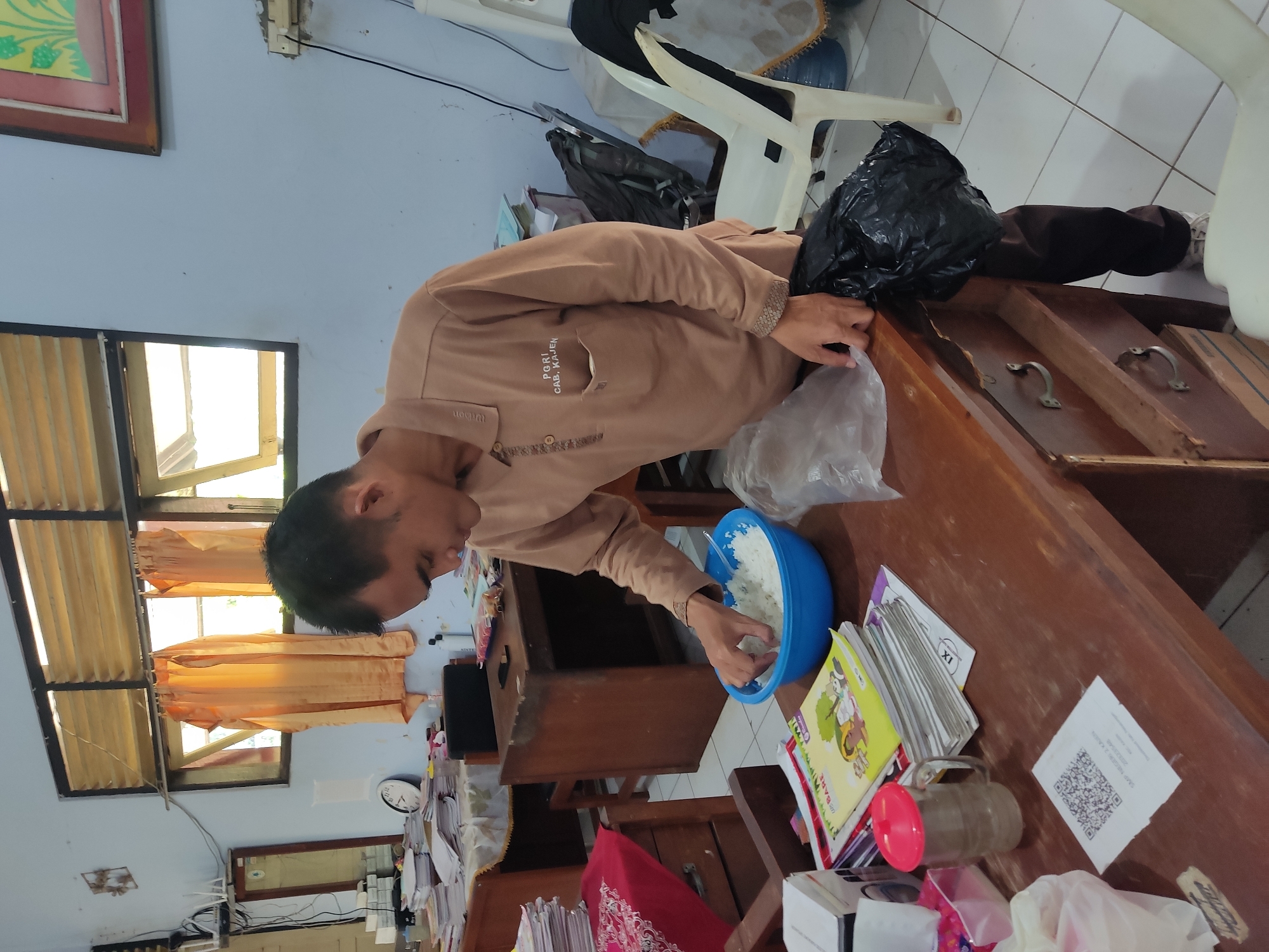 In an indoor classroom setting, a man wearing a light brown polo shirt with the insignia "PGRI CAB. KAJEN" is standing at a wooden desk. He is holding a plastic bag in his right hand and reaching into a blue bowl filled with white rice with his left hand, appearing to be serving himself or preparing food. The desk is cluttered with various items, including stacks of books and papers, a glass mug with a red lid, a QR code printed on a white slip of paper, and a black plastic bag. In the background, there are wooden desks, chairs, a mirror, a clock, and windows with orange curtains. The room has light blue walls and a tiled floor.