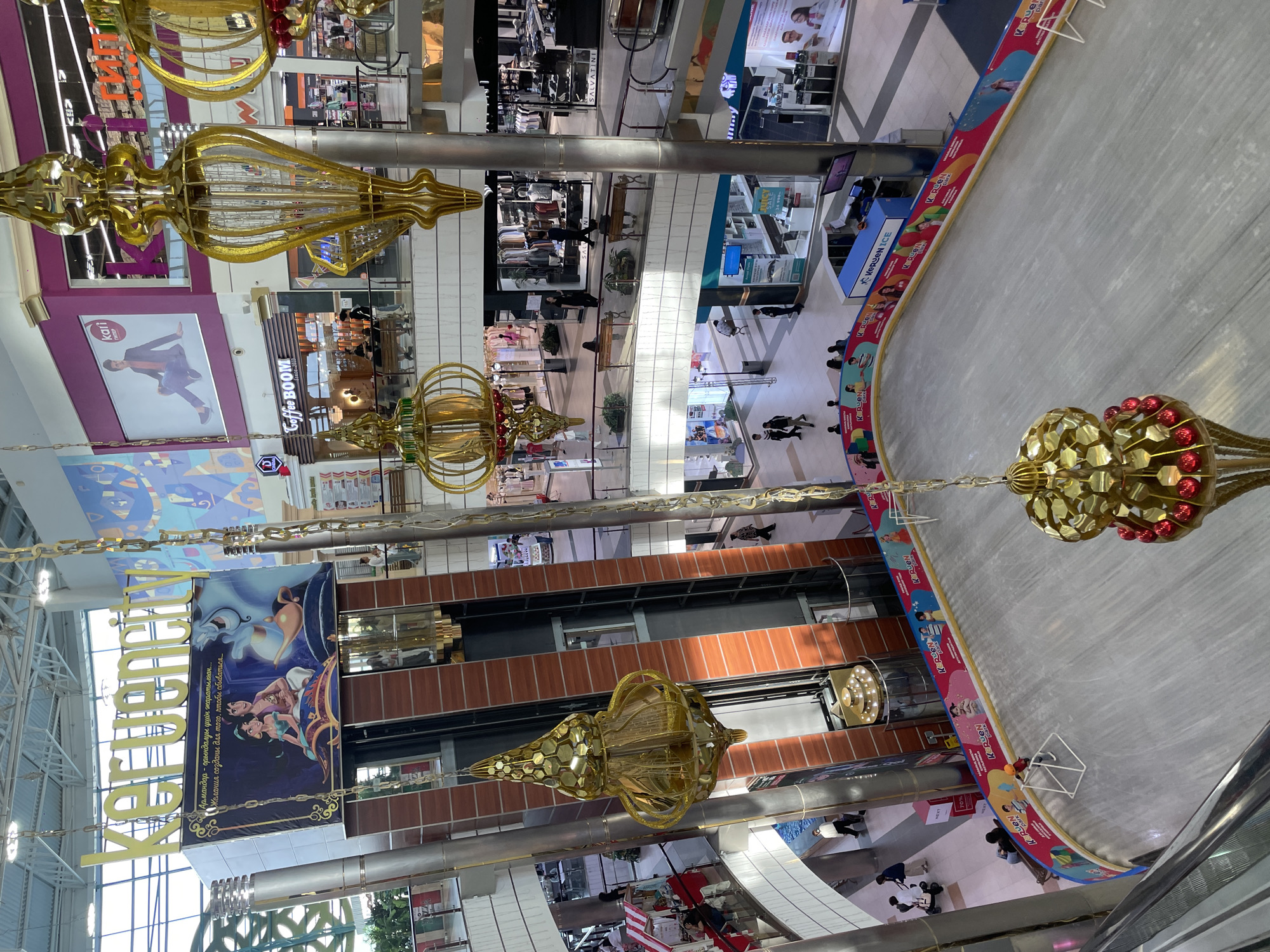 The image captures an indoor view of the K-ruu en City shopping mall in Aktobe, Kazakhstan, a multi-level complex featuring an ice skating rink in the foreground. The rink is surrounded by a colorful banner adorned with cartoon characters and the mall's logo, "K-ruu en ICE." A few individuals, including a child wearing a helmet, are present on the ice. Suspended from the ceiling are large, ornate golden lanterns, contributing to the mall's festive atmosphere. Various shops and escalators are visible on the upper levels, with shoppers moving about. A prominent advertisement featuring characters from Disney's Aladdin is displayed near the mall's entrance. The overall environment appears to be a lively and modern commercial space.