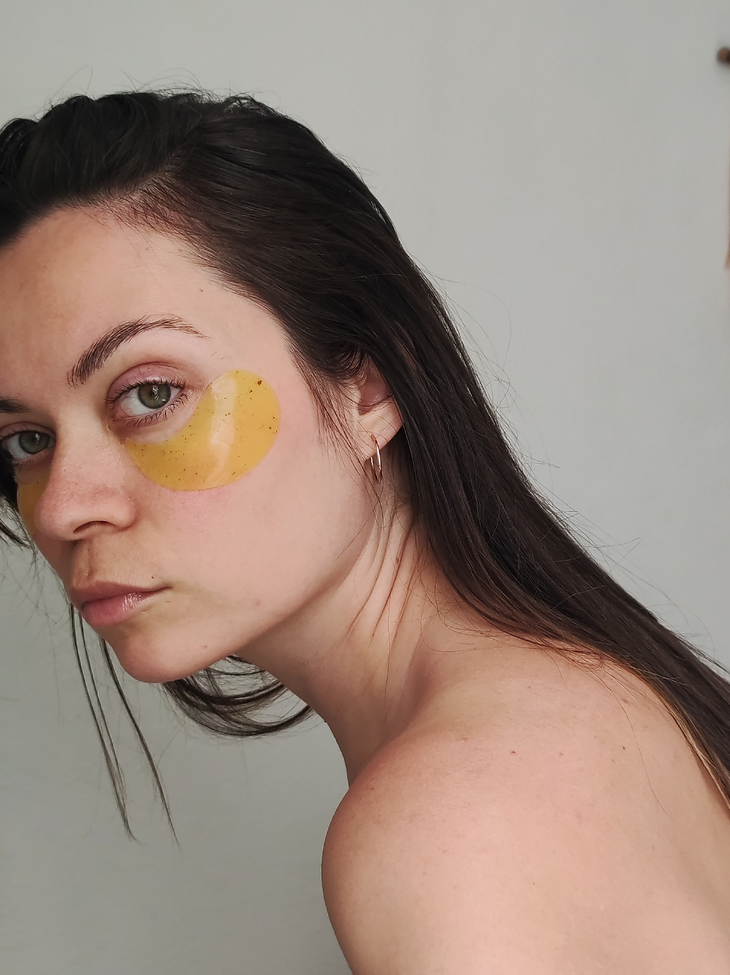 A close-up studio portrait of a fair-skinned woman with long, dark brown hair. She has light green eyes and is wearing a golden-yellow under-eye patch on each cheek. A small gold hoop earring is visible on her left ear. The background is a neutral, light gray. The image does not contain information regarding Chaikovsky, Russia.