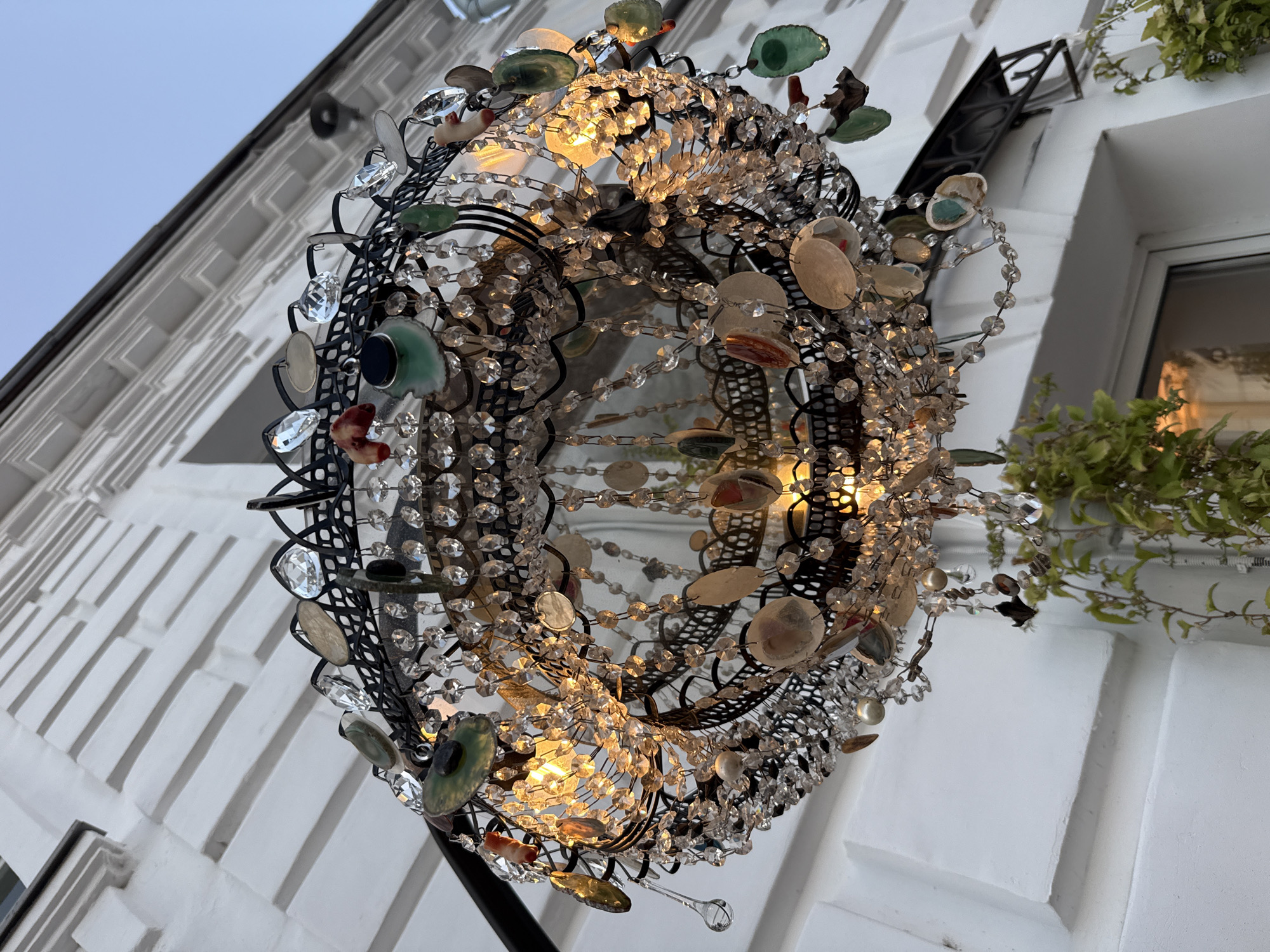 A close-up, low-angle shot captures an ornate chandelier hanging in front of a white, textured building. The chandelier is a circular, multi-layered structure made of black metal, adorned with numerous clear crystals, semi-precious stones such as agate slices and jade, and shell fragments. The stones and crystals hang at various lengths, creating a dense, sparkling effect. Several small lights are visible within the chandelier, casting a warm glow. Green vines trail down from the top right of the frame, partially obscuring the building and the lower portion of the chandelier. The building's facade features a decorative, stepped pattern and appears to be an older architectural style. The sky above is a pale blue, suggesting either dawn or dusk. The scene appears to be an exterior decoration for a shop or establishment in Zhukovo, Russia.