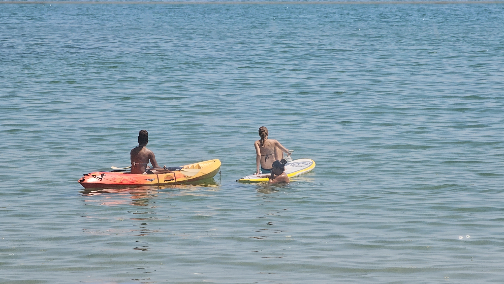 The image shows a sunny day at the Black Sea coast in Varna, Bulgaria. Three people are in the water. On the left, a person is sitting in an orange and yellow kayak. To the right of the kayak, another person is sitting on a yellow paddleboard. Next to the paddleboard, a third person is partially submerged in the water. The water is calm with gentle ripples, and the sky is clear. The scene appears to be a leisurely activity on a summer day.