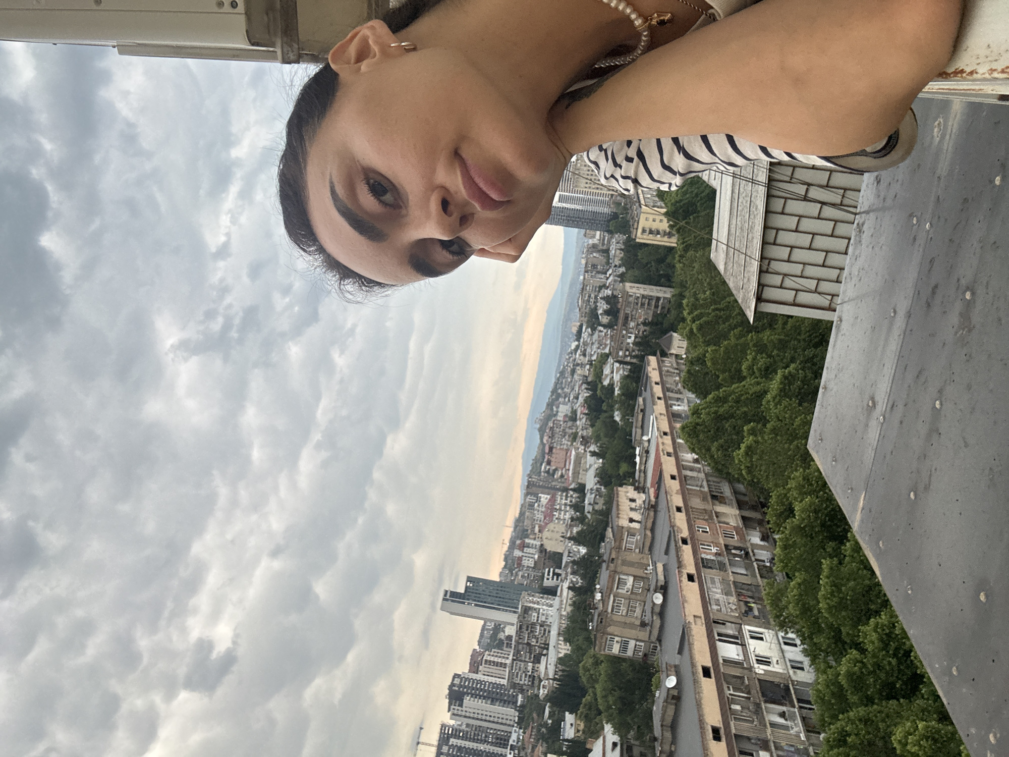 A close-up, over-the-shoulder selfie features a young woman leaning her head on her hand, looking directly at the camera. She has dark hair pulled back and is wearing a striped shirt and a pearl necklace. She is on a balcony, with a city skyline in the background. The city appears to be Tbilisi, Georgia, based on the architecture and urban landscape. The buildings range from older, apartment-style blocks to more modern high-rises, interspersed with lush green trees. The sky is overcast with a muted, cloudy atmosphere. The foreground of the image shows parts of the balcony railing and rooftop.