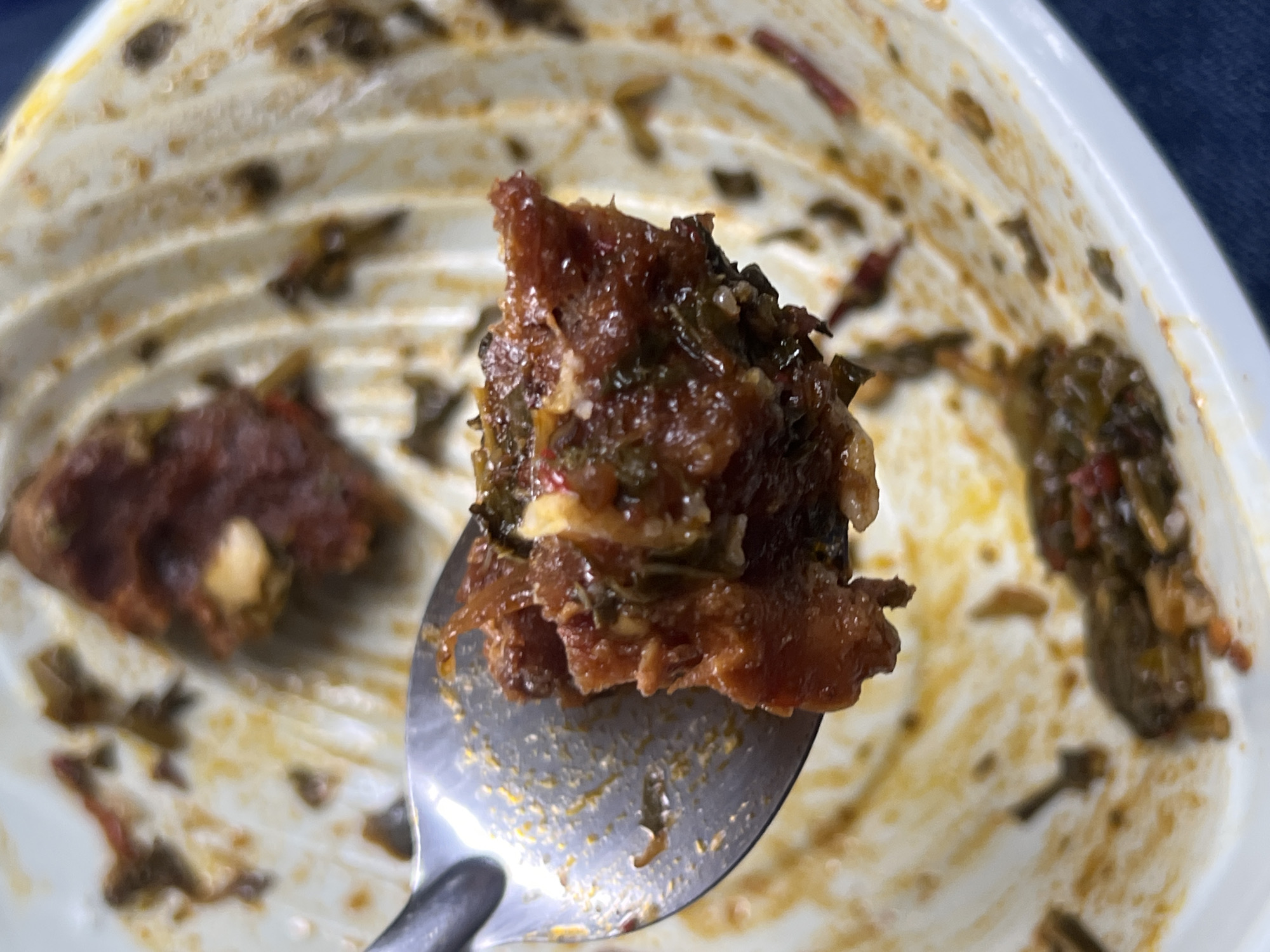 A close-up shot captures a spoonful of dark, reddish-brown food being held above a white, ribbed container. The food appears to be a stew or mixed dish, with discernible green leafy elements and lighter, possibly creamy or cheesy, specks integrated throughout. The metal spoon holding the food is positioned in the foreground, angled slightly downwards. In the background, the white container shows remnants of the same food adhering to its interior, along with yellowish-orange stains from the meal. The container's ribbed texture is prominent. The scene depicts the act of eating, likely a casual meal. No people, visible text, time of day, weather, or specific emotions are visible. This meal is situated in Jalingo, Nigeria.