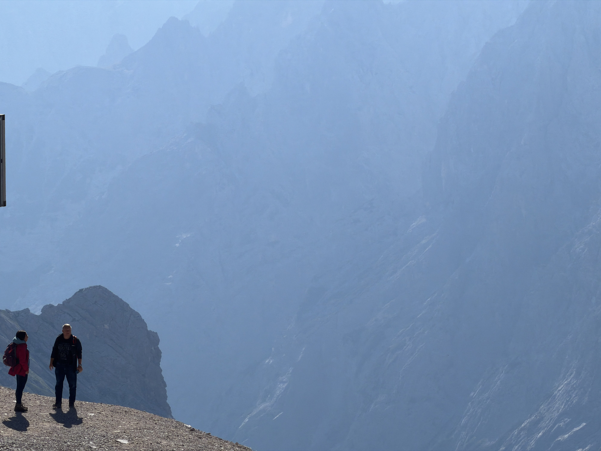 This media file captures a breathtaking mountain landscape, likely in Garmisch-Partenkirchen, Germany, under a clear, bright sky, suggesting midday. Two individuals are positioned on a rocky outcrop in the foreground, appearing to be hikers.
The person on the left is wearing a red backpack and a dark jacket over a light-colored top. They are looking towards the person on the right. The second individual, a man, is dressed in a dark, long-sleeved shirt with visible text and dark pants. He is looking to his left, towards the mountain range. Both are standing on a gravelly path, casting distinct shadows, indicating a sunny day.
The backdrop is dominated by a vast, hazy expanse of mountains. The layers of peaks fade into a soft blue haze, creating a sense of immense depth and scale. The textures of the distant mountains are visible, with rugged slopes and shadowed crevices, hinting at a challenging terrain. A prominent, sharp peak is visible directly behind the two hikers, adding to the dramatic scenery.
There is a partial view of a white signpost on the far left edge of the frame, its purpose or text not discernible. The overall mood is one of awe and exploration, with the vastness of nature and the presence of hikers suggesting an outdoor adventure. The clear visibility of the foreground and the hazy, distant mountains indicate a significant elevation and possibly a clear atmospheric condition that allows for such dramatic depth perception.