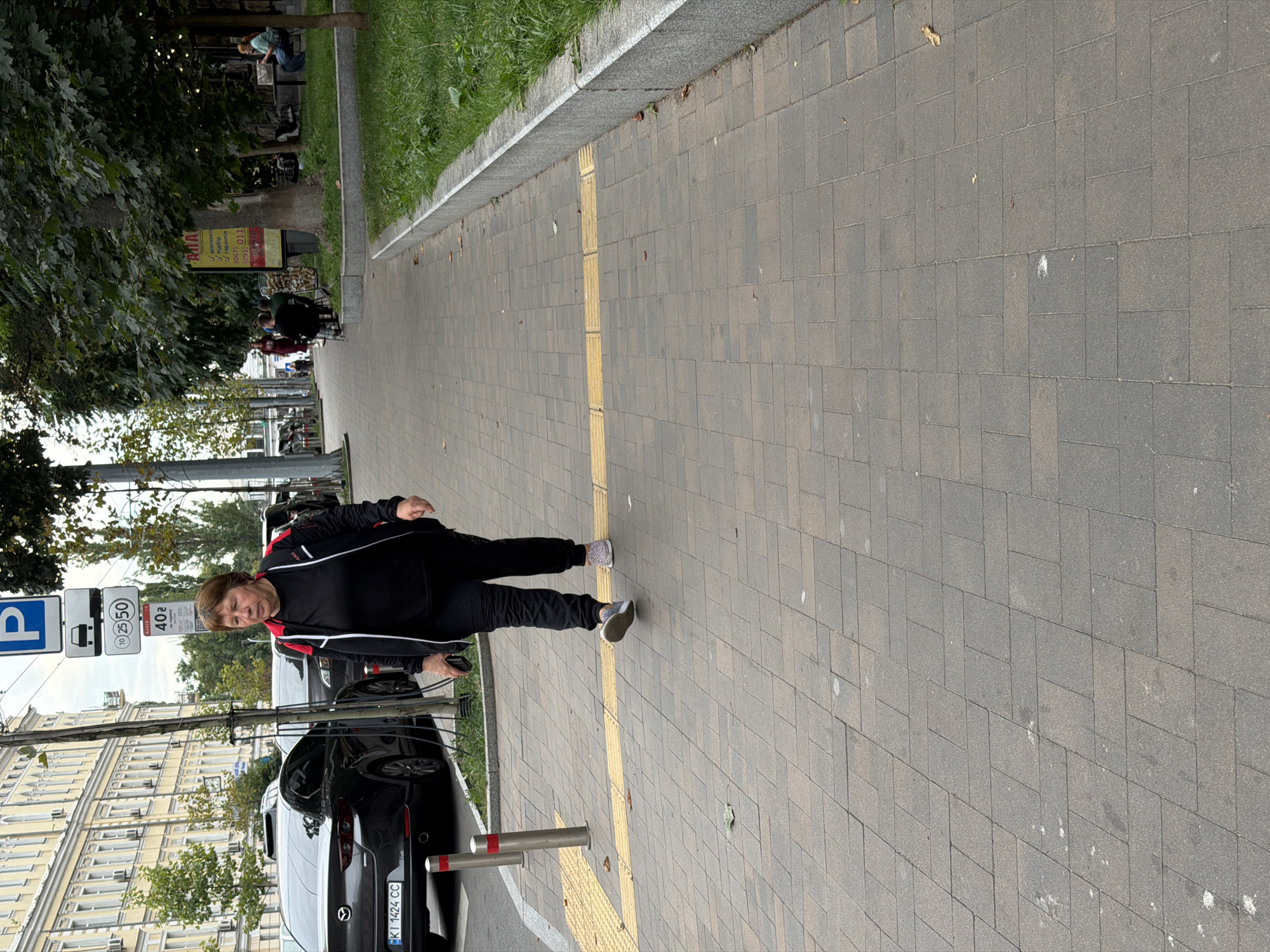 The image shows a woman walking on a sidewalk in Kyiv, Ukraine. She is wearing black athletic pants and a black jacket with red accents, and she is holding a smartphone in her right hand. The sidewalk is paved with gray rectangular stones and has a yellow tactile paving strip running along its edge. To the left of the sidewalk are parked cars and a building with a light-colored facade. To the right of the sidewalk is a patch of green grass, trees, and a cafe with outdoor seating. Several road signs are visible in the background, including a parking sign and speed limit signs.