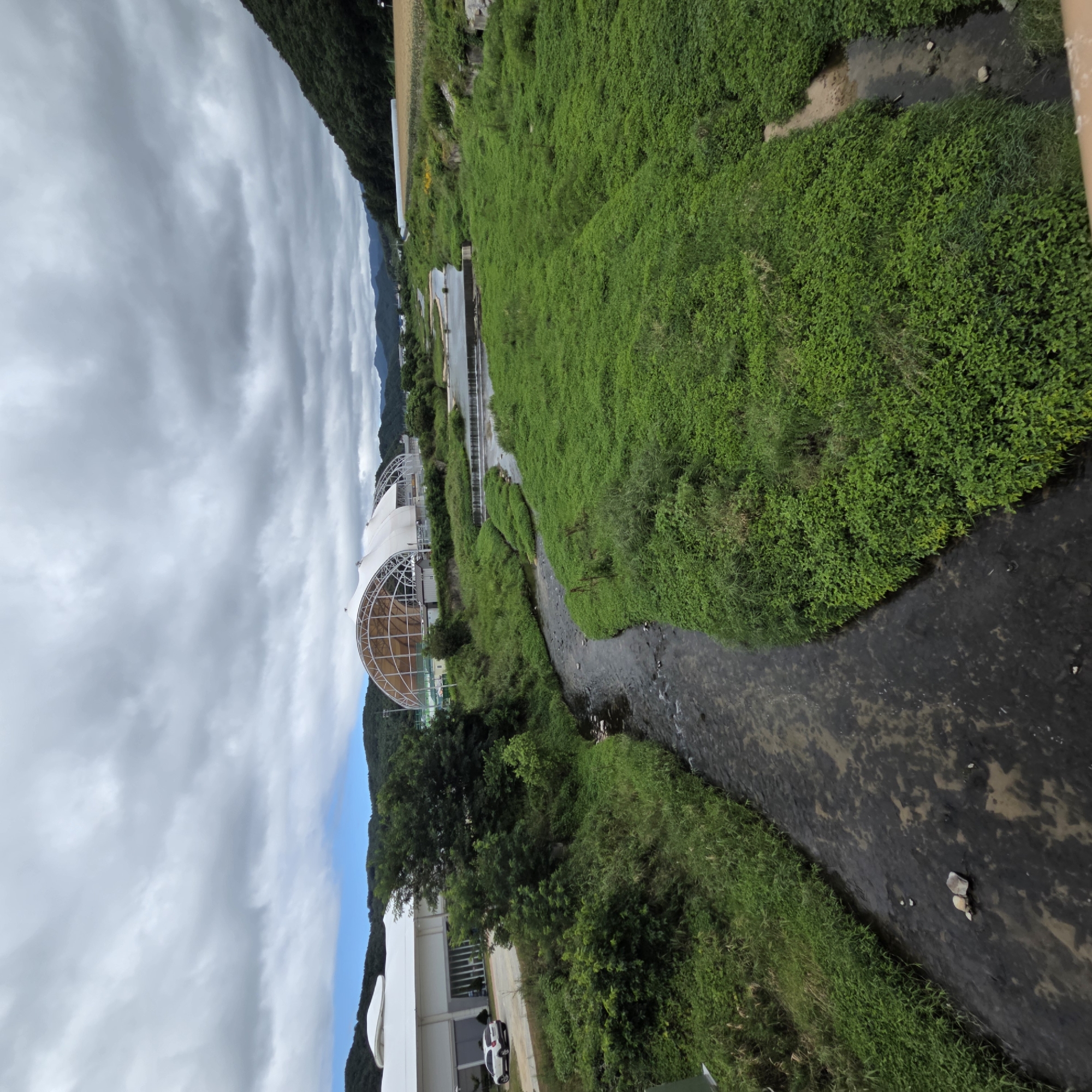 The image, taken in Dunnae-myeon, South Korea, captures a natural landscape with a river flowing through it. The river meanders through lush green vegetation, with the banks densely covered in plants. In the background, a large, modern building with a distinctive arched roof structure is visible. The sky is overcast with heavy clouds. Hills covered in trees form the backdrop on the right side of the image. The scene appears to be a serene, outdoor environment, likely a park or recreational area.