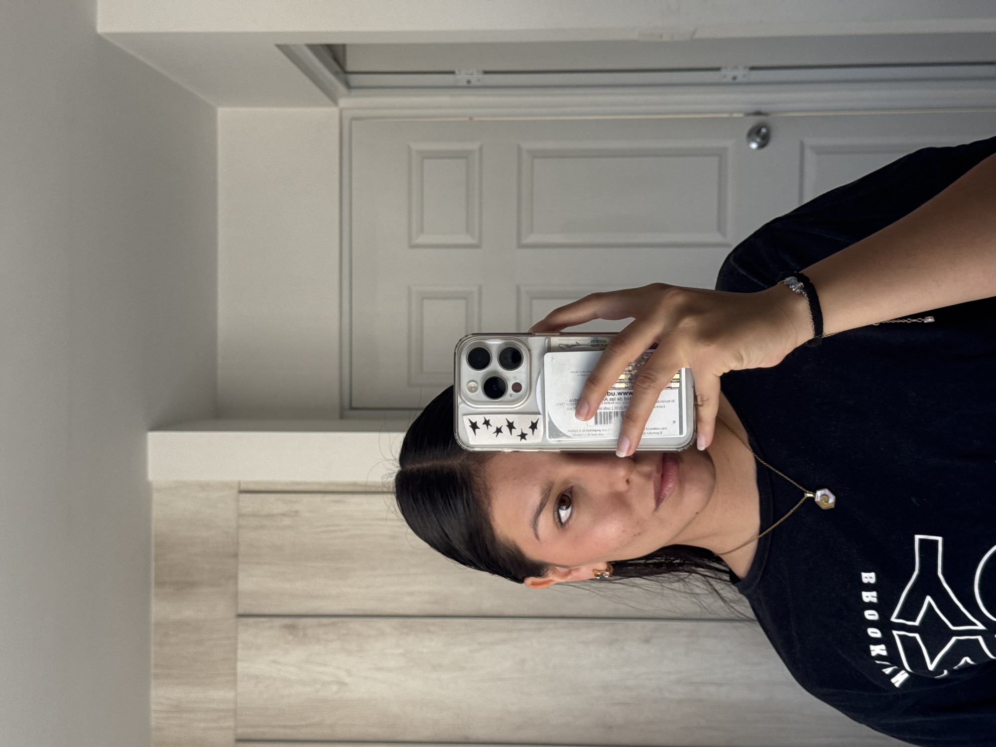 A woman with dark hair is taking a mirror selfie indoors. She is holding an iPhone with a clear case decorated with black stars. She is wearing a black t-shirt with the word "BROOKLYN" partially visible in white letters and a gold necklace. Behind her is a white door with three recessed panels and a silver doorknob, and to the left is a light-colored wall and what appears to be a light-colored vanity. It is not possible to confirm the location of San Francisco Ocotlán (Ocotlán), Mexico from this image.