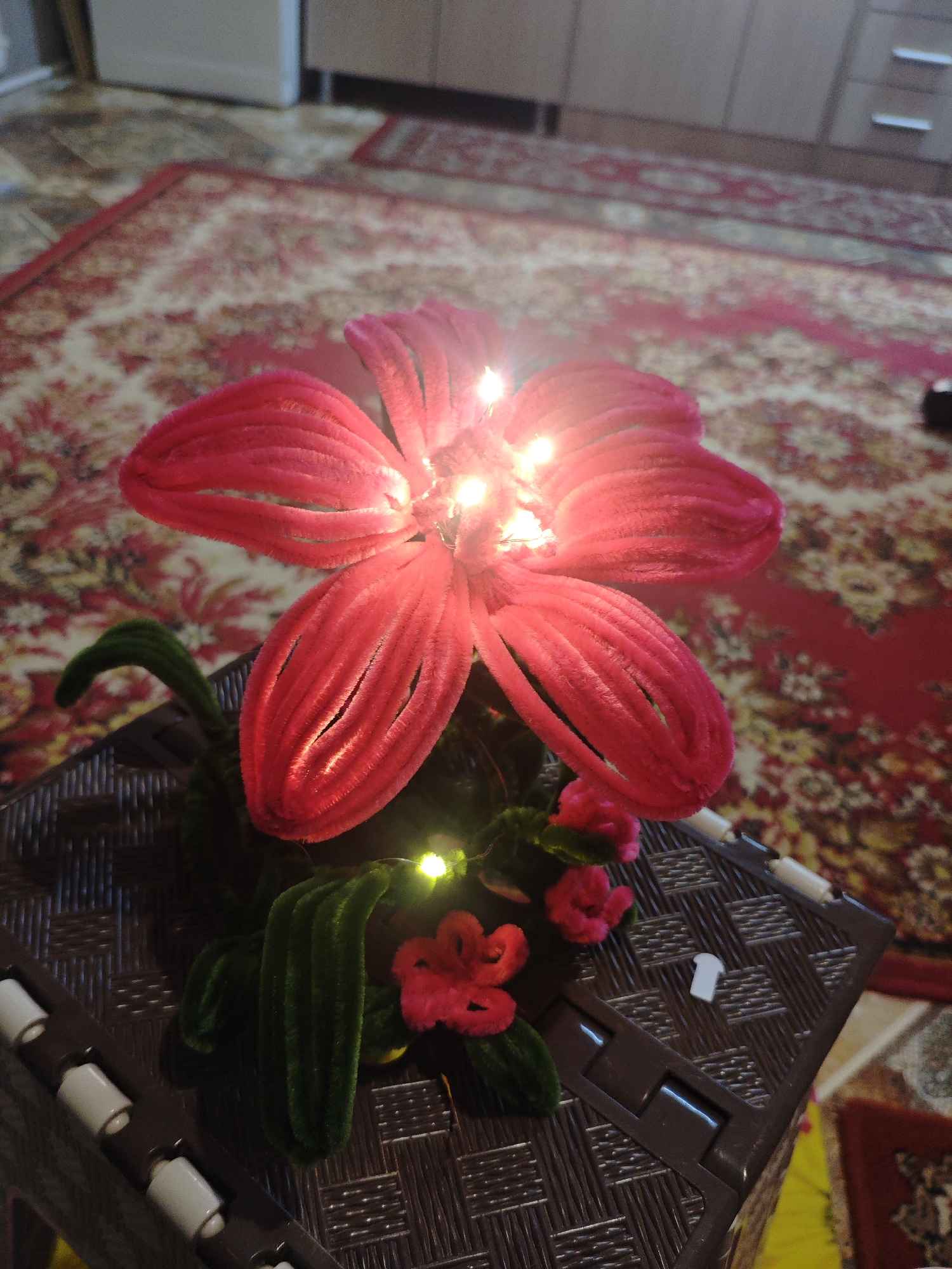 A bright red, pipe cleaner flower with small lights embedded in its center rests on a brown, woven plastic surface. The flower has five large petals, each creased lengthwise. Green pipe cleaner leaves extend from the base of the flower, with two smaller red pipe cleaner flowers also present. The object is positioned on a brown plastic container with a textured surface, featuring white cylindrical accents along its side and a brown latch. In the background, a red and cream patterned rug is visible, with a section of what appears to be a tiled floor to the left. The lighting suggests an indoor setting. The image was taken in Armavir, Russia.