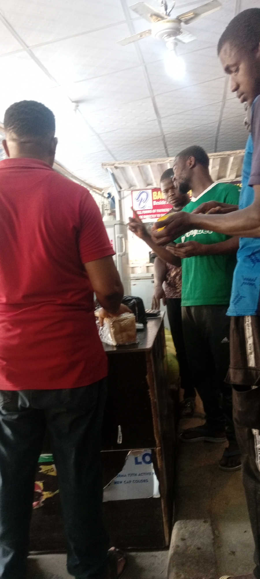 In Aviation Quarters, Nigeria, the scene depicts a group of people gathered at a counter. A person in a red shirt stands with their back to the viewer, cutting a loaf of bread on the counter. Three other individuals stand to the right, appearing to be receiving slices of mango. One man wears a green shirt, and another wears a blue shirt. The setting seems to be a small shop or eatery with a ceiling fan overhead. A sign with the letters "LO" is visible on the front of the counter.