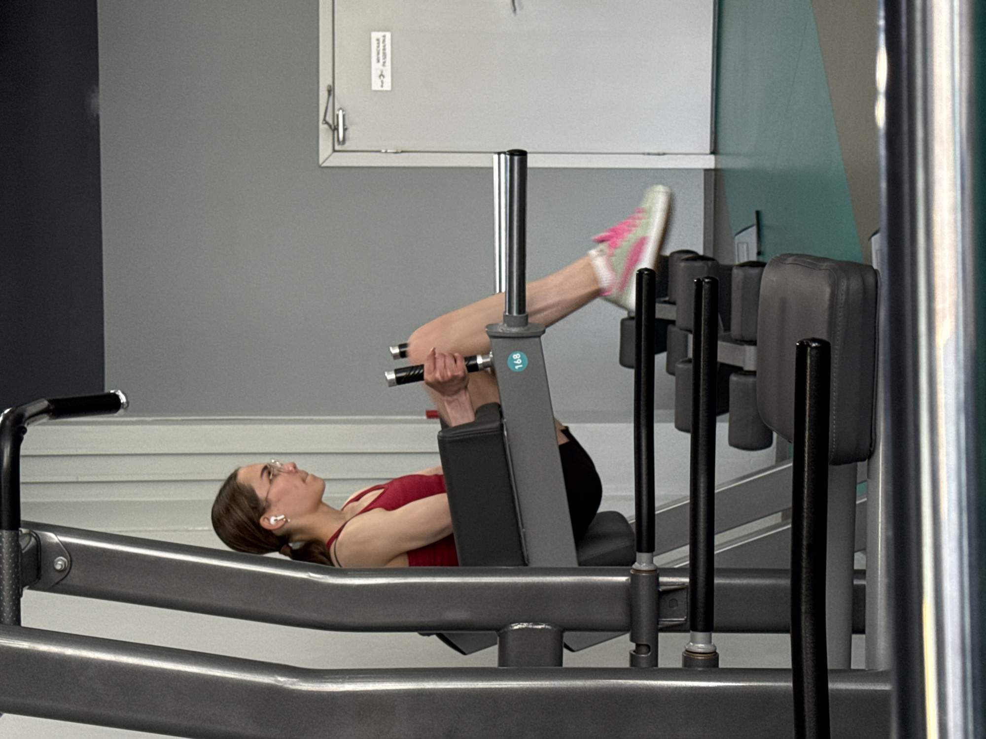 A woman is using a leg press machine in a gym. She is lying on her back with her legs extended and pressing against the padded platform. She is wearing a red tank top, black pants, eyeglasses, and white sneakers with pink accents. She has earphones in her ears. The machine is made of gray metal and has black padding. The background includes a gray wall, a white door with a metal latch, and a pale green wall panel. The location is a gym, but there is no evidence to suggest this is in Yekaterinburg, Russia.