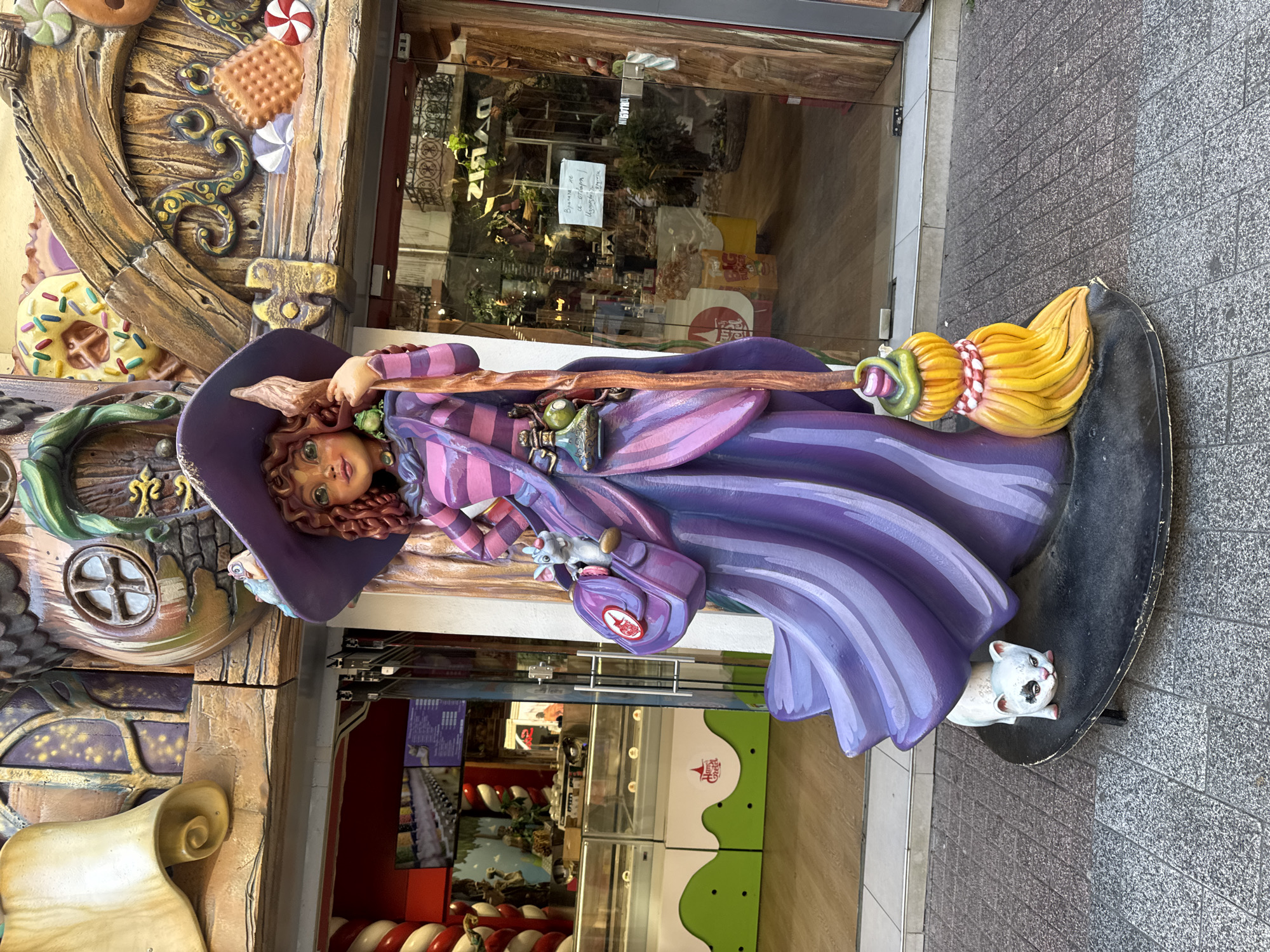 This image captures a whimsical, life-sized statue of a witch standing on a paved sidewalk in front of a building. The witch is depicted with curly reddish-brown hair, green eyes, and a friendly expression. She wears a purple hat and a striped purple and pink dress, holding a broom in her right hand and a satchel with a small mouse on her left hip. A small white cat with a black patch over its eye is posed at the base of the statue. The building behind the statue appears to be a shop, with a facade decorated with candy-themed elements like donuts, cookies, and lollipops. The entrance features glass doors and windows, revealing displays of goods inside, including what appears to be an ice cream parlor on the left. The statue and building suggest a playful, perhaps Halloween-themed, establishment. This scene is located on "The Street of Crafts" in the Old Town of Plovdiv, Bulgaria.