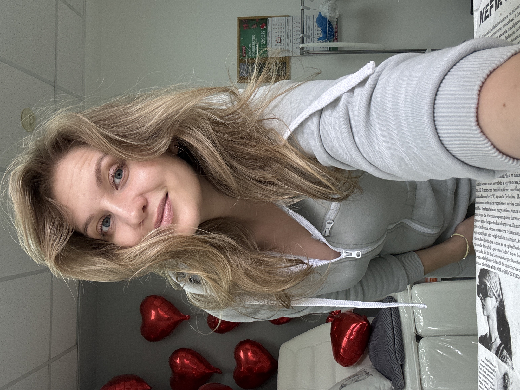 A fair-skinned woman with blonde hair and blue eyes, wearing a light grey hoodie, is the primary subject. She is positioned at an angle, with red heart-shaped balloons visible in the lower part of the frame, suggesting a celebratory or romantic theme. A calendar indicating the year 2026 is visible in the background. The woman is indoors, possibly in an office or administrative space, suggested by the ceiling tiles and shelving in the background. Text in both Russian and Spanish is also visible, possibly on printed materials or packaging. The location within Izhevsk, Russia, cannot be determined from the visual information provided.