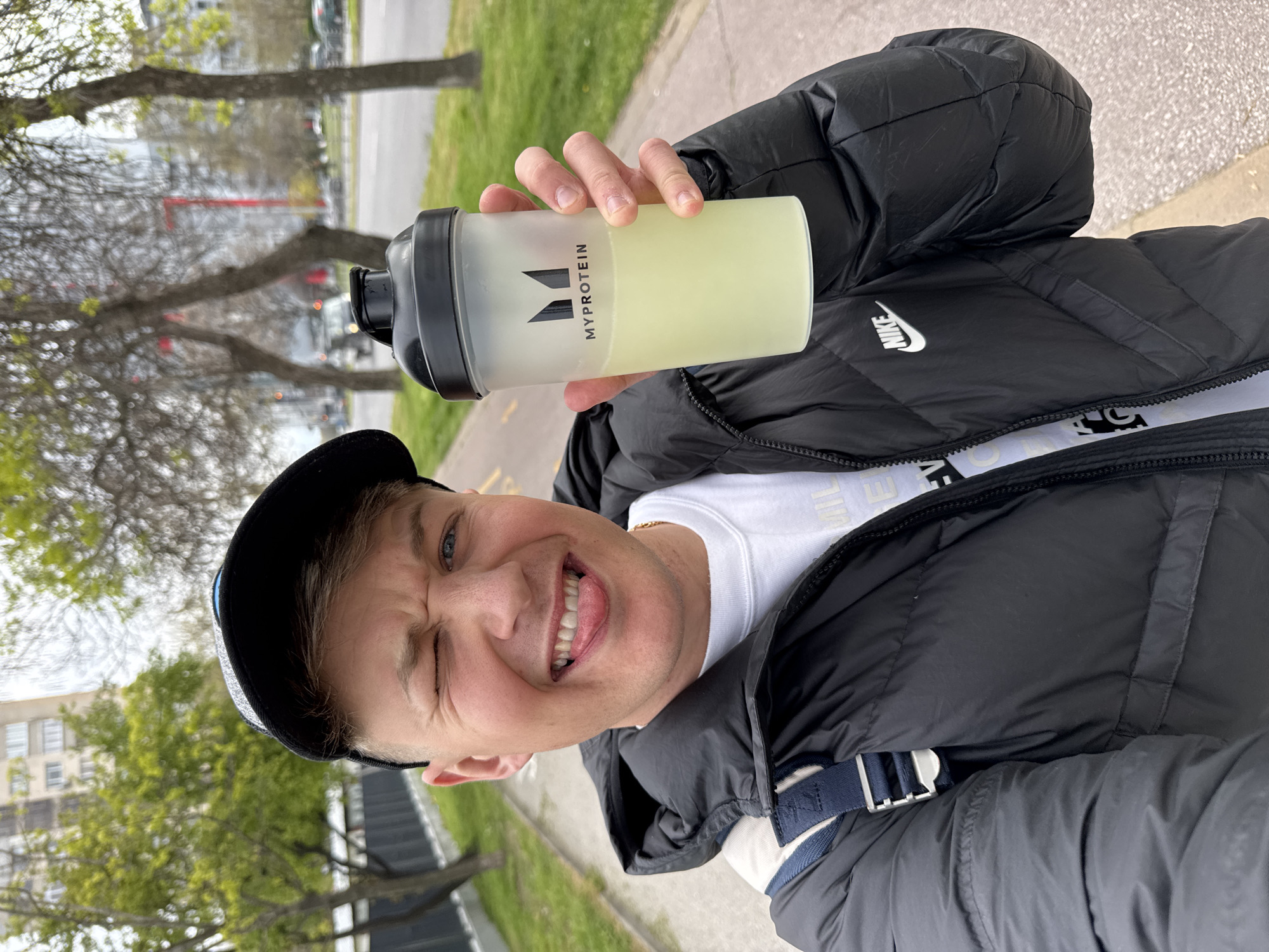 A close-up, eye-level shot shows a man holding up a "MyProtein" shaker. He is positioned on an outdoor walkway next to a street, likely in a city. The man wears a black baseball cap and a black Nike puffer jacket over a white shirt. He winks and sticks out his tongue. Buildings and bare trees are visible in the background. It is not possible to ascertain if the location is the city of Plovdiv, Bulgaria.