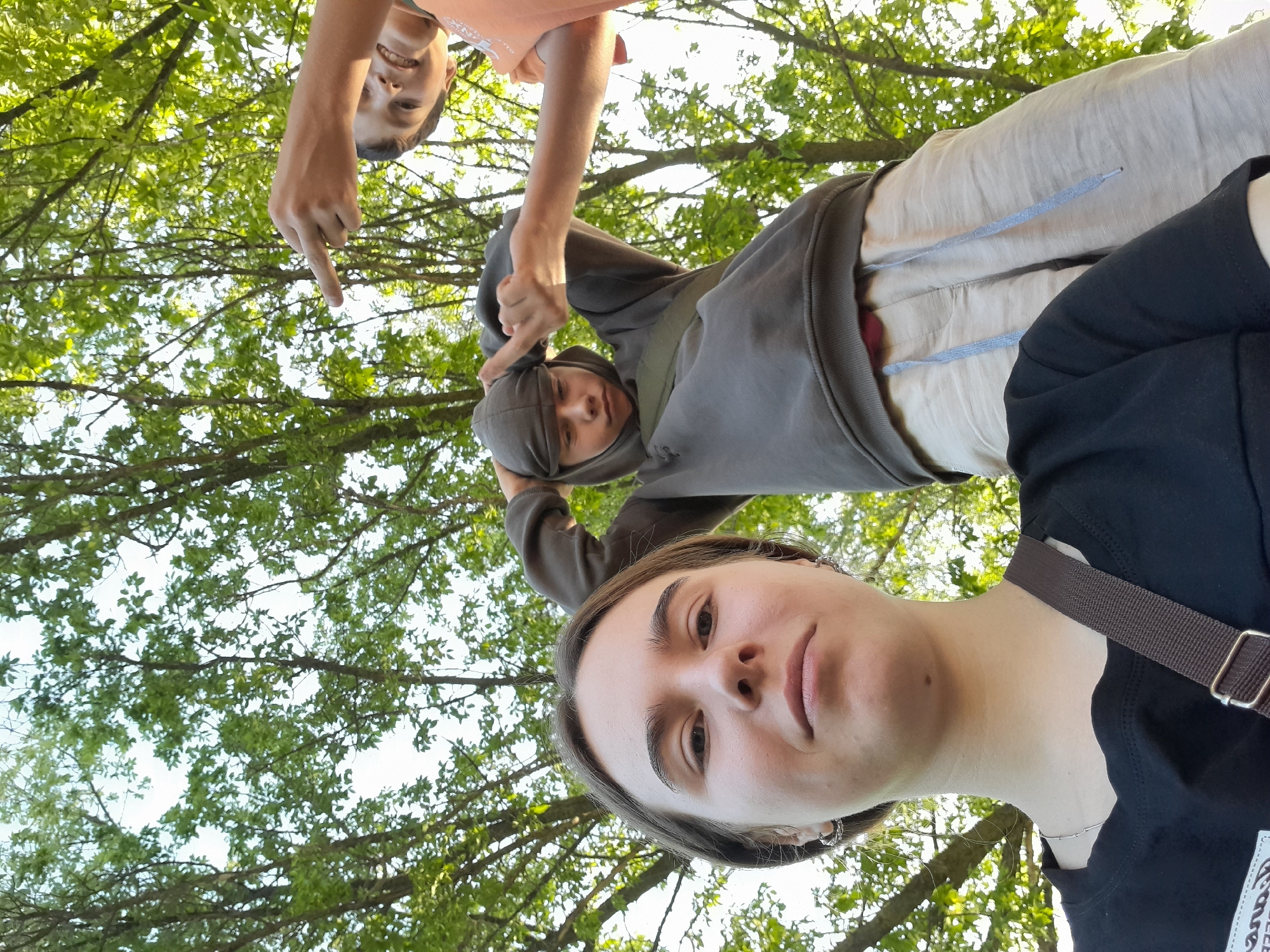 Three young individuals are outdoors, with a low-angle perspective looking up at them against a backdrop of lush green trees and a bright sky. In the foreground, a person with light brown hair, wearing a black t-shirt and hoop earrings, looks directly at the camera with a neutral expression. To their right and slightly behind, another person in a grey hooded sweatshirt and light-colored shorts is visible, with their hood pulled up and hands near their head, as if adjusting it. To the right of the hooded figure, a third person wearing an orange t-shirt is partially visible, smiling and pointing their index finger towards the left, seemingly at the other individuals. The overall impression is a candid, informal snapshot taken in a natural, outdoor setting.