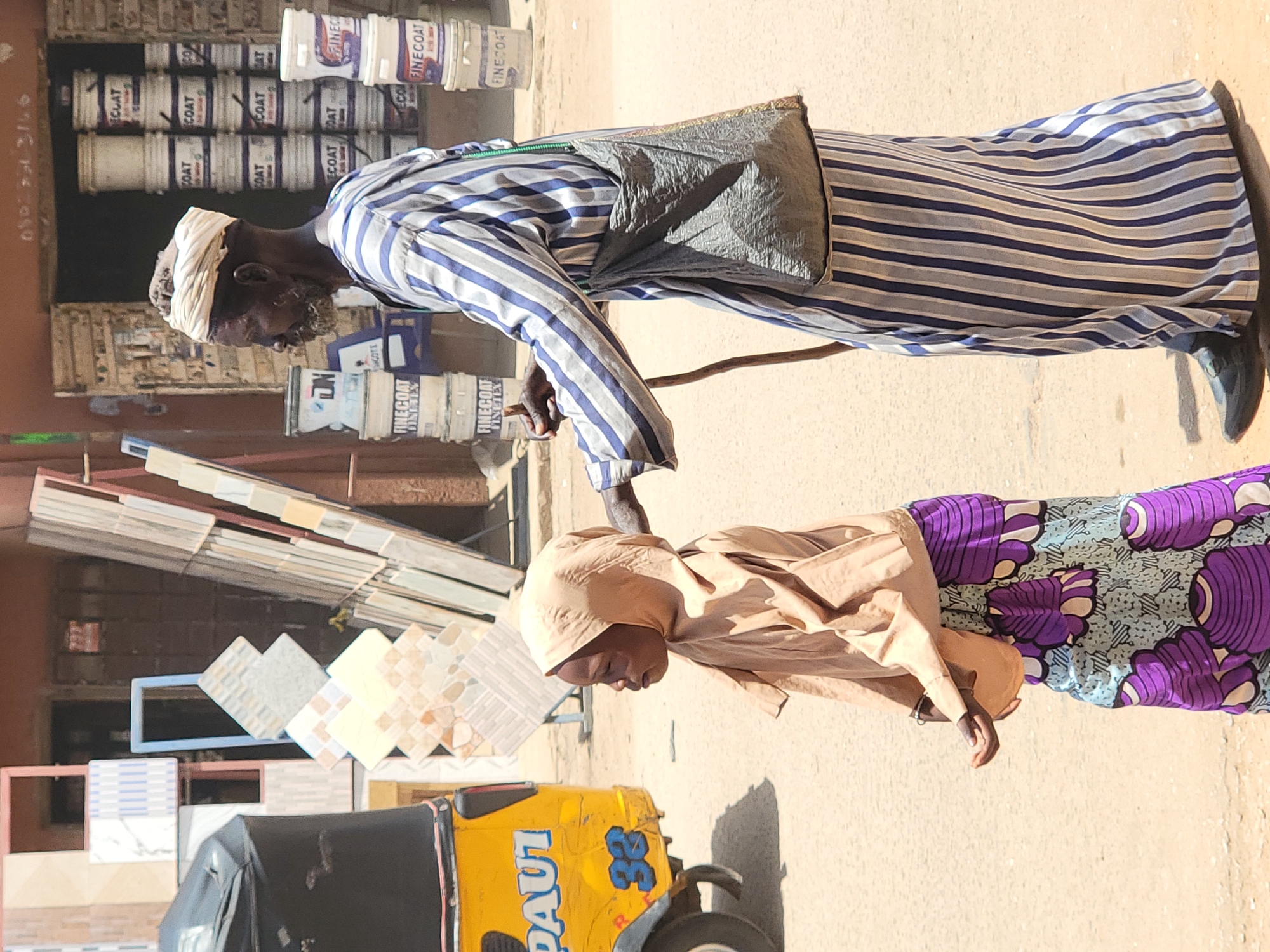 Here's a detailed description of the image:
The photograph captures a scene on a street in what appears to be Maiburiji, Nigeria. The image is dominated by two individuals: an elderly man and a young child.
The elderly man is wearing a long, striped blue and white robe with a gray satchel slung across his shoulder. He has a white turban on his head and carries a walking stick. He is bent over, seemingly directing the child.
The young child is clad in a light brown robe and a vibrant purple and blue patterned skirt. The child's head is covered with a hood.
In the background, there's a display of building materials, including tiles and buckets of paint labeled "FINECOAT FINETEX." To the left is a yellow and black tricycle taxi, with the word "DAUT" and the number "32" visible on its side.
The scene is brightly lit, indicating it is daytime, and the setting appears to be a bustling commercial area.