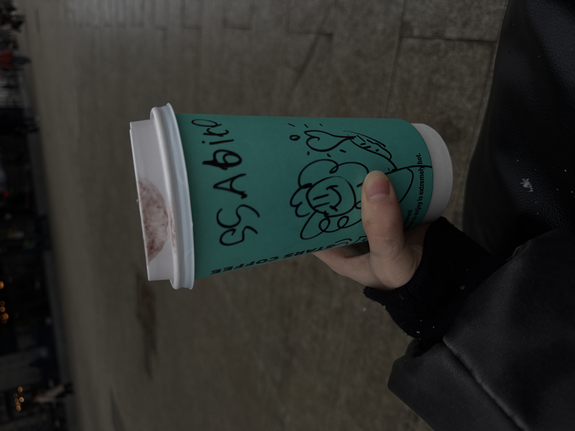 A close-up, tilted shot depicts a hand holding a turquoise paper coffee cup with a white lid. The cup is labeled "STARS COFFEE" vertically in black marker, alongside a whimsical illustration of a smiling face with flower petals and hearts. The warning "be careful to enjoy is extremely hot" is partially visible near the cup's upper edge. The hand is wearing a black sleeve and a black leather jacket. The background shows an out-of-focus urban setting with buildings, possibly indicating a city. Based on the "STARS COFFEE" branding and the style of coffee cup, the location could be inferred as Moscow, Russia, where the Stars Coffee chain operates. The ground appears to be paved, and the lighting suggests daytime, possibly overcast conditions.