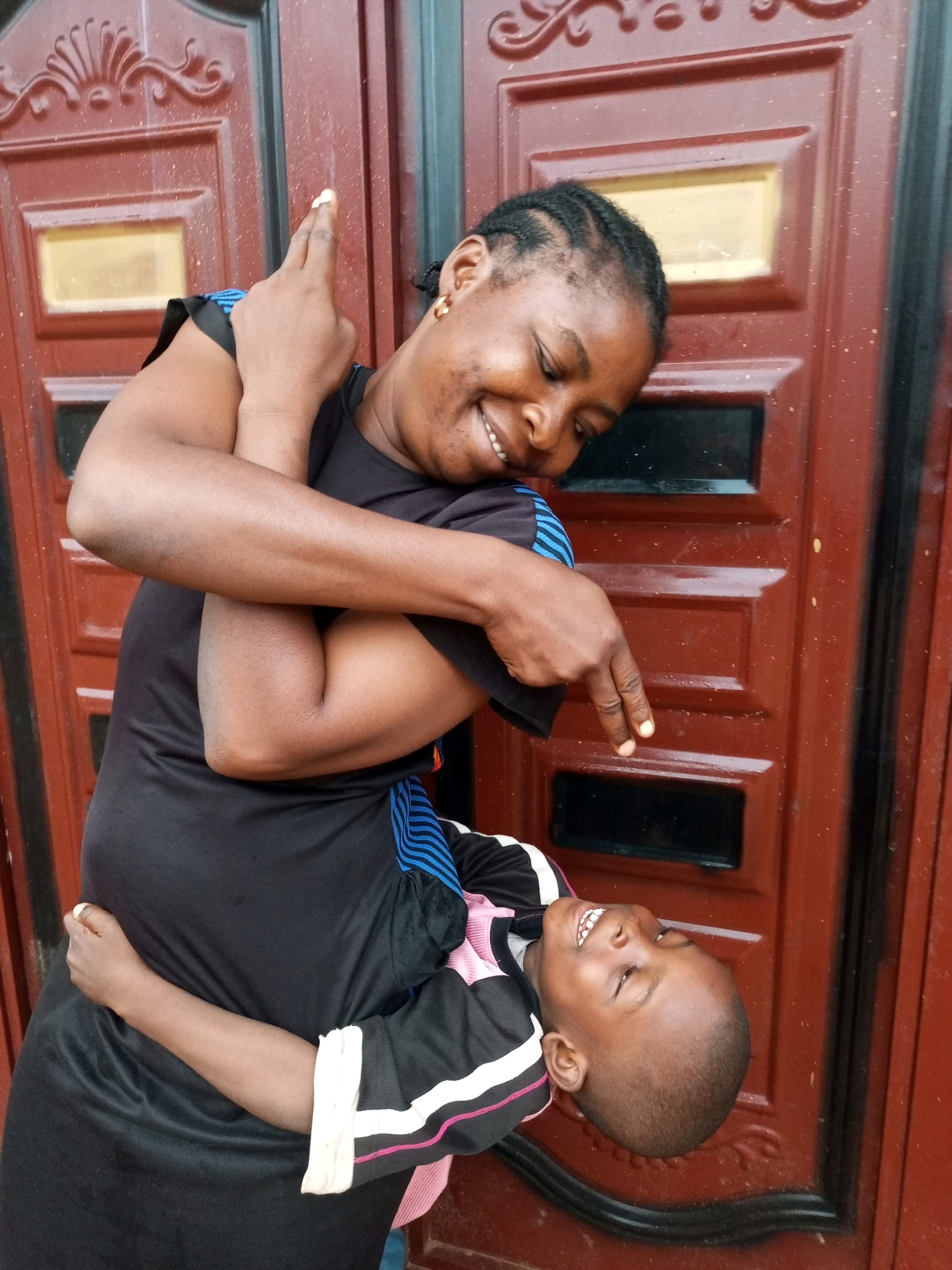 In Tempure, Nigeria, a woman with braided hair and gold hoop earrings embraces a child in front of a reddish-brown door with black accents. The woman is wearing a black t-shirt with blue stripes on the sleeves, and the child is wearing a black shirt with white and pink stripes. The woman is looking down and smiling, while the child is looking up and laughing. The woman's left arm is around the child's back, and her right arm is raised, with her index and middle fingers extended. The child's arms are wrapped around the woman's neck. The door appears to be made of metal and has decorative elements. The scene is outdoors, and the lighting suggests it is daytime.