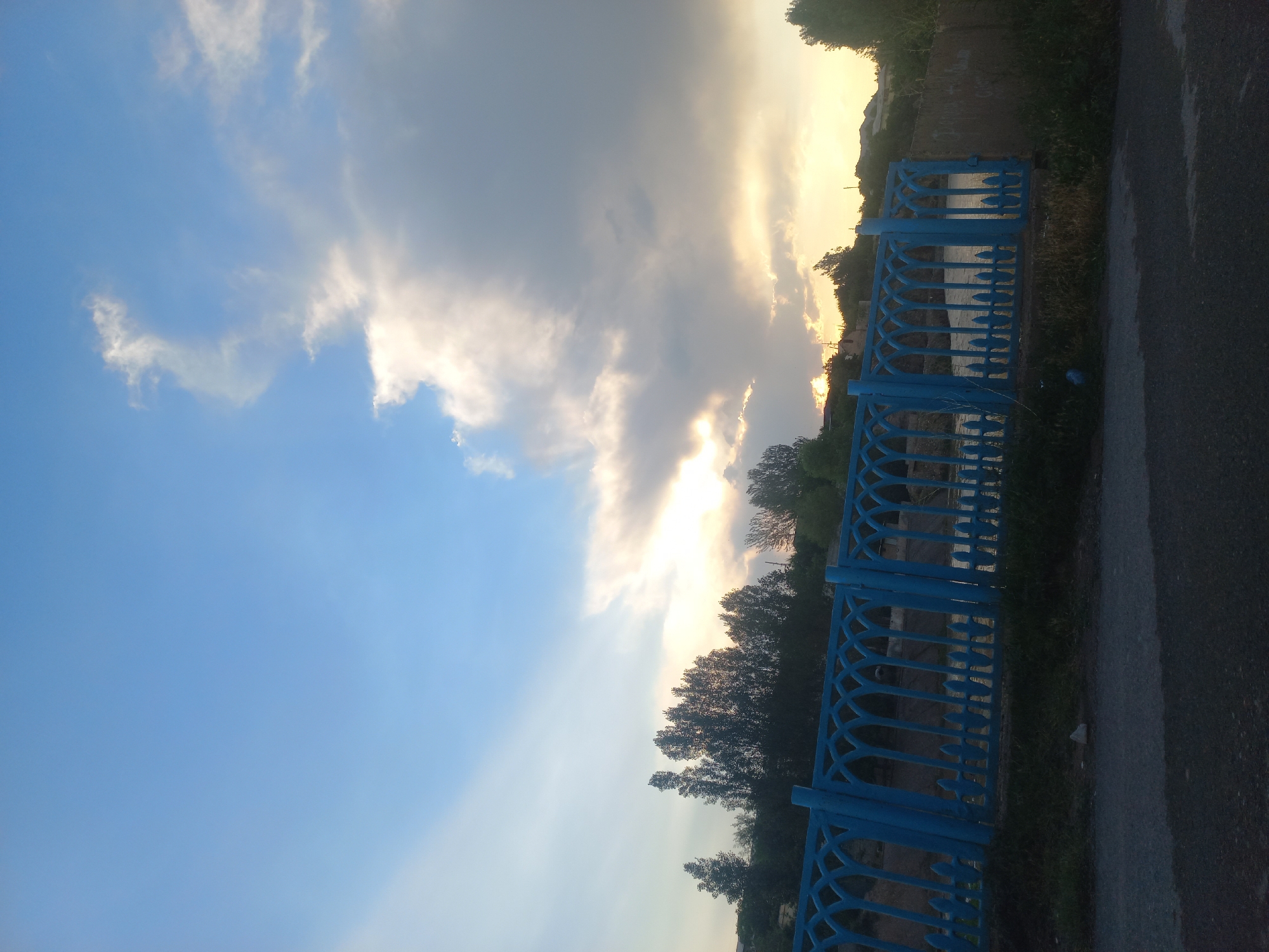 The image captures a serene outdoor scene, likely during sunset, given the warm glow of the sun breaking through dramatic clouds. The sky is a dominant feature, transitioning from a clear, bright blue on the left to a mix of grey and golden-tinged clouds on the right. The sun's rays are visible, creating a luminous effect.
In the foreground, a bright blue metal fence with an intricate, decorative pattern runs horizontally across the frame, suggesting it might be a railing or barrier along a path or waterfront. Beyond the fence, a body of water is visible, reflecting the sky. The scene is bordered by lush greenery and trees, which are silhouetted against the bright sky, particularly on the right side of the image. A paved road or pathway runs alongside the fence on the far right, appearing somewhat wet, possibly indicating recent rain or dew.
There are no people or specific objects identifiable that indicate an activity or event. The overall mood is peaceful and tranquil, enhanced by the natural beauty of the sky and the surrounding landscape. The location cues of Kasansay, Uzbekistan, might suggest a rural or semi-rural setting with natural waterways and vegetation. No visible text is present in the image.