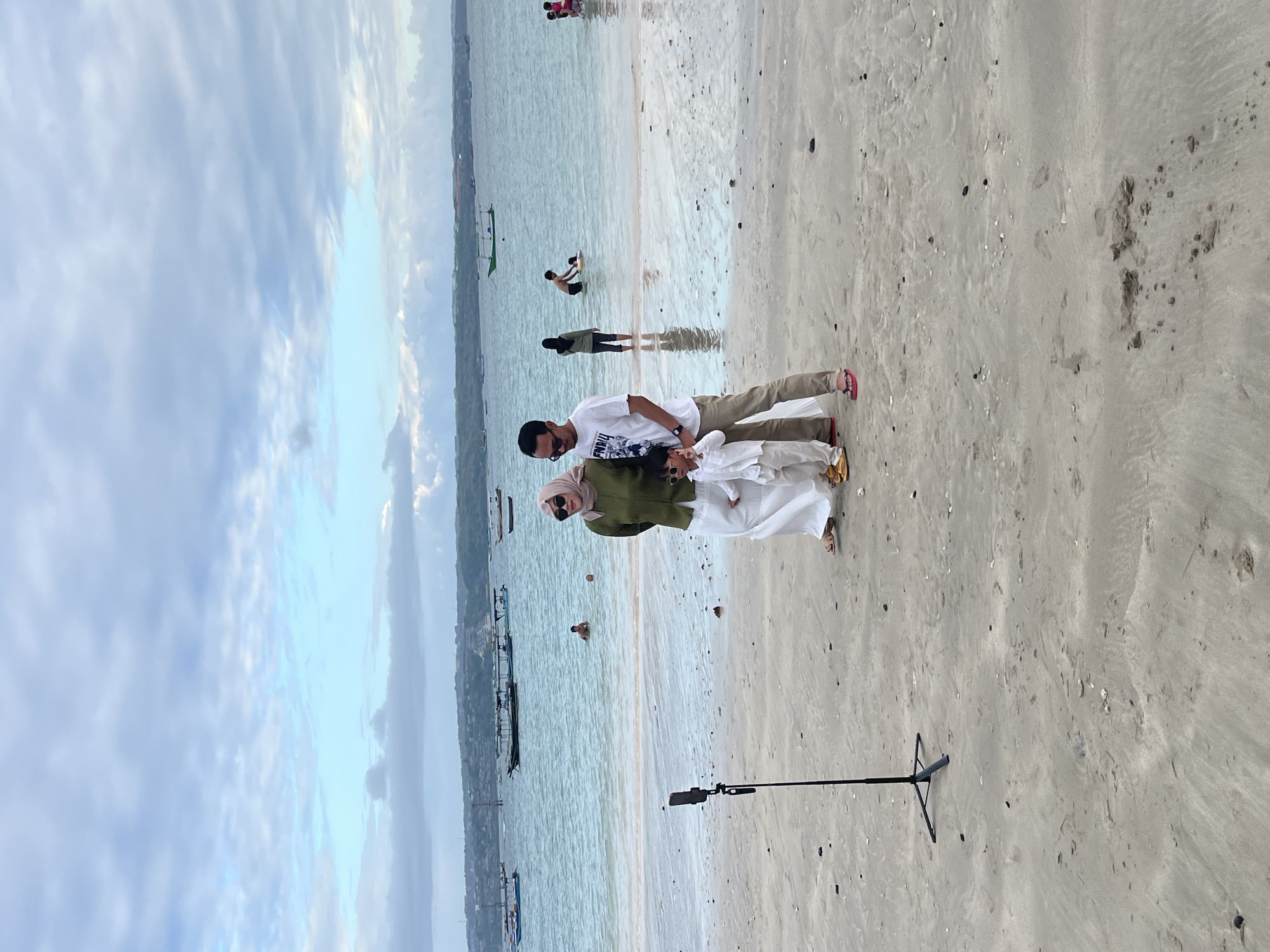 The image depicts a family posing for a photo on a beach in Kuta, Indonesia. A father, mother, and young child are standing on the wet sand near the waterline. The mother wears a light pink hijab and a green long-sleeved top with a white skirt. The father is dressed in a white t-shirt and khaki pants, and the child is in a white outfit. A smartphone on a tripod is positioned in front of them, suggesting they are taking a selfie or recording a video. In the background, the ocean stretches out with several boats anchored offshore and some individuals swimming or wading in the water. The sky is cloudy, with patches of blue visible. The beach itself is sandy with scattered small stones and debris.
