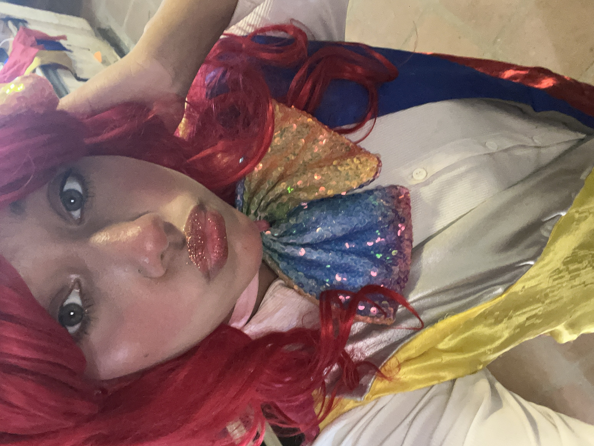 A close-up, angled shot shows a person with red hair and glittery red lipstick. The person has fair skin and gray eyes. They are wearing a multicolored sequined bow. Behind them is a white shirt with a blue, red, and yellow cloth draped over it. The location is unknown.