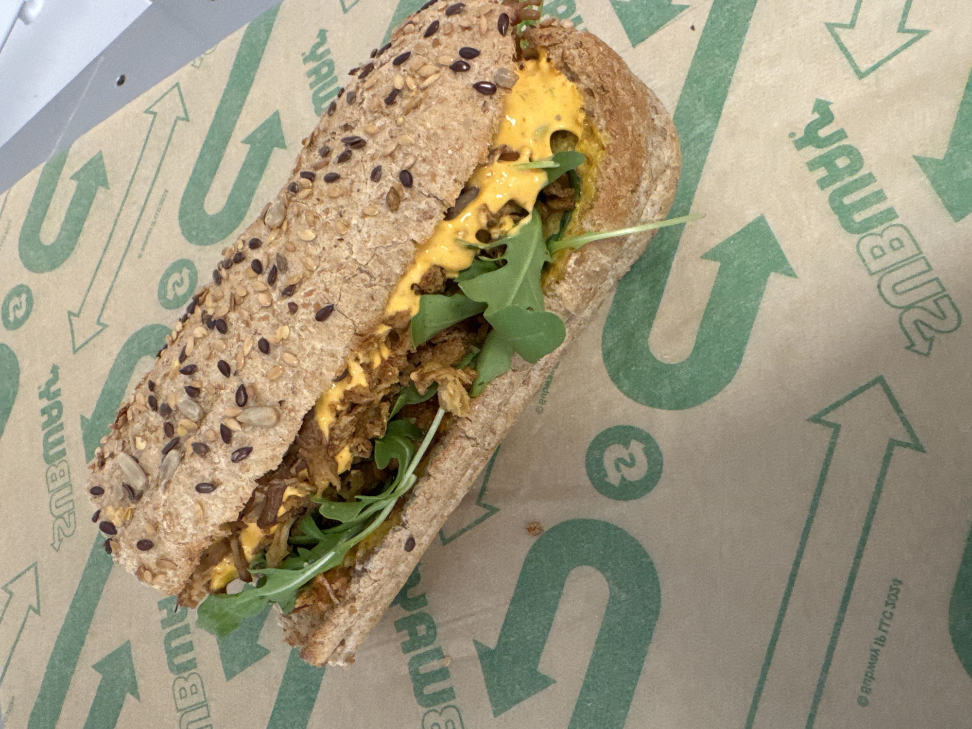 A close-up, overhead shot shows a fully assembled sandwich on a patterned paper wrapper. The sandwich is made with a whole wheat bun topped with various seeds, including sunflower and black sesame seeds. Inside the bun, visible through a cut down the middle, is shredded meat, green arugula leaves, and a bright yellow sauce. The wrapper beneath the sandwich features repeating green arrows forming circular patterns and the word "SUBWAY" reversed. The lighting is overhead and even, with no shadows cast. The background is a light brown color, consistent with the wrapper. The location is listed as Unknown, Unknown.
