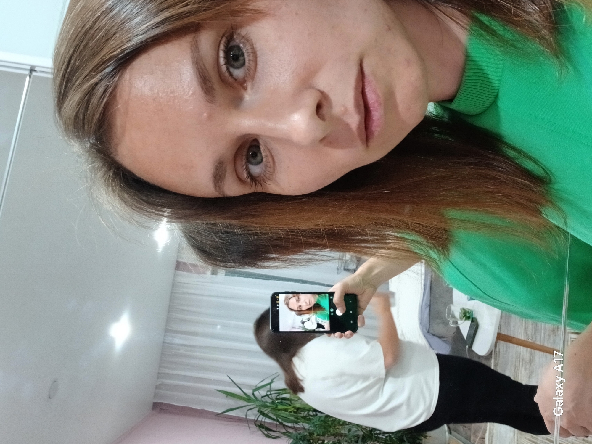 A woman is taking a selfie in a room. She is wearing a green top and has light brown hair. She is holding a black smartphone with the screen showing a reflection of her taking the picture. In the background, there is another woman wearing a white top and black pants, along with a plant and a table. The room has white walls and curtains, and the lighting appears to be soft and natural. There is no information to discern the specific location in Yekaterinburg, Russia.