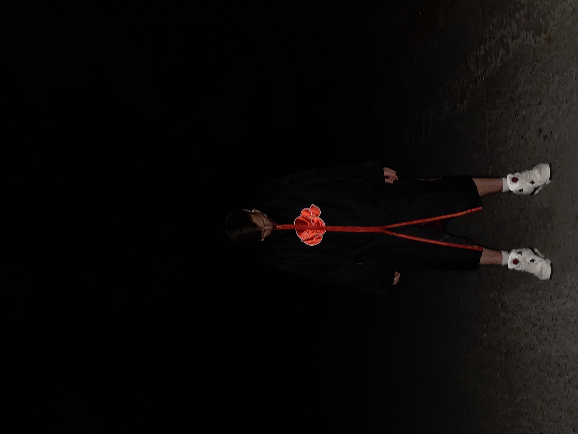 A man in an Akatsuki cloak lies on a dark pavement. The only visible light reflects off his white sneakers and the pavement in the upper right of the frame. The location is unknown, and there is no indication that this image was taken in Lyaskovo, Bulgaria.