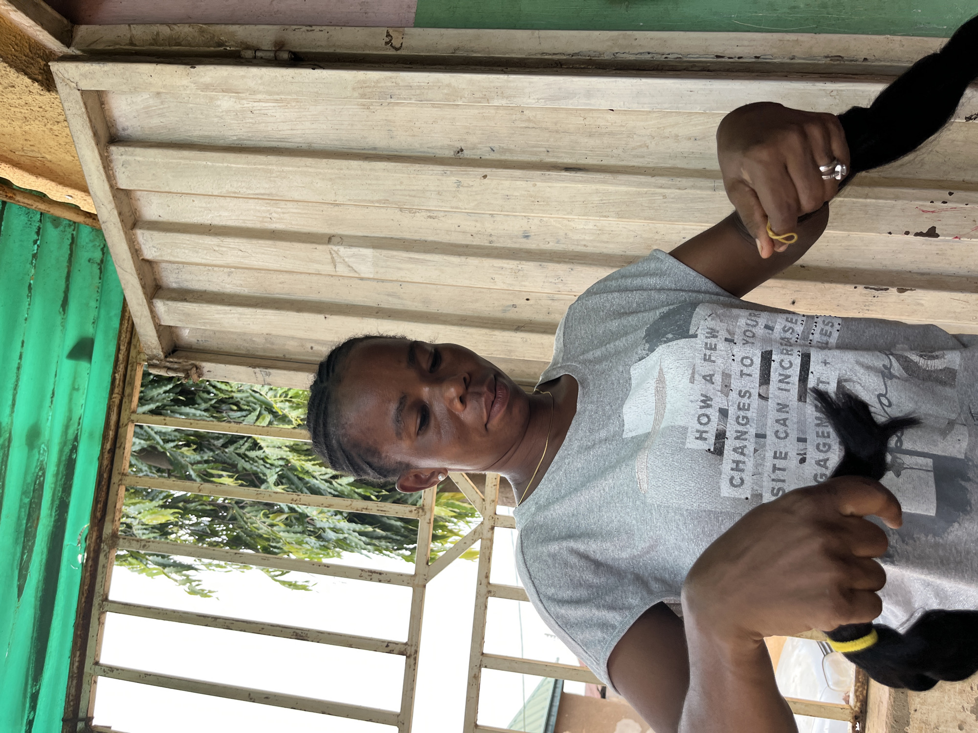 A medium, indoor, eye-level shot shows an African woman with braids seated and wearing a gray t-shirt with words on it, holding a black bunch of hair tied with a yellow rubber band in each hand; she has a gold necklace on, is wearing a ring, and is looking at the camera. Behind her is a structure with gray, horizontal planks. Through an iron-barred opening, there are green trees and other structures. The image was taken in Madalla, Nigeria.