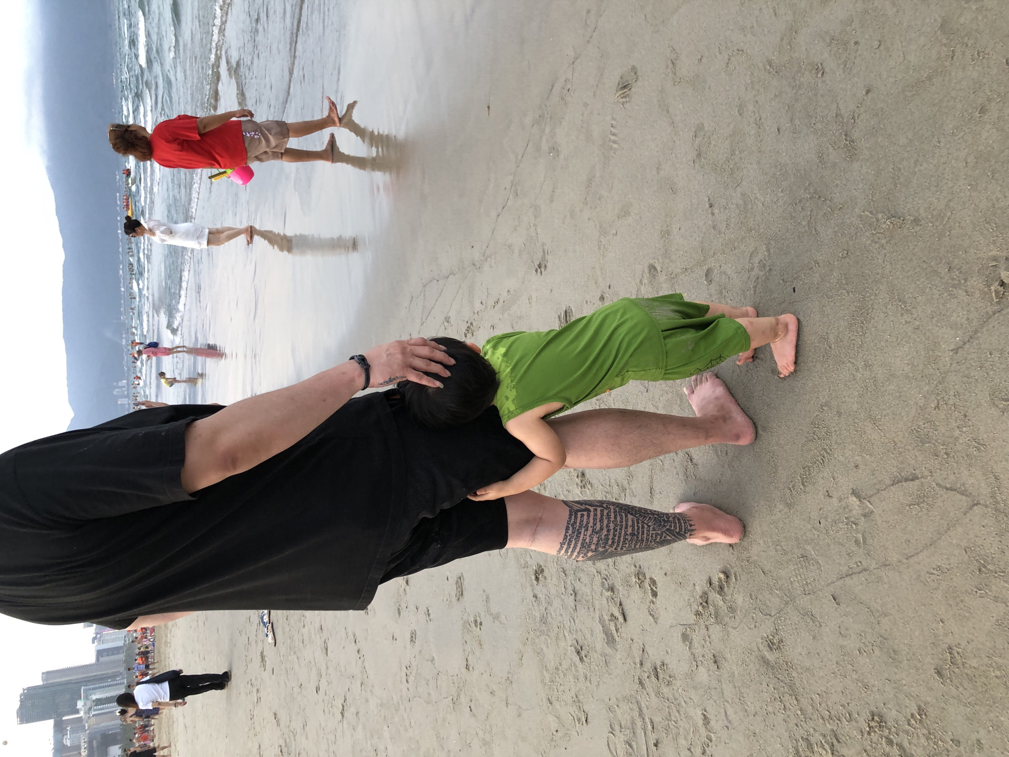 The image depicts a beach scene in Đà Nẵng, Vietnam, likely during the day. In the foreground, a person wearing a black t-shirt and shorts is partially visible, with their arm around a small child dressed in a green outfit. The child is leaning against the person's chest, with their head resting on the person's shoulder. The person has a large tattoo on their leg. The beach is sandy, with some footprints visible. In the background, the ocean is visible with waves, and several other people are scattered along the shoreline. Further back, there are buildings, suggesting an urban area adjacent to the beach. The weather appears to be overcast or hazy, with a soft light illuminating the scene. The overall mood seems peaceful and relaxed, with the focus on the interaction between the two individuals.