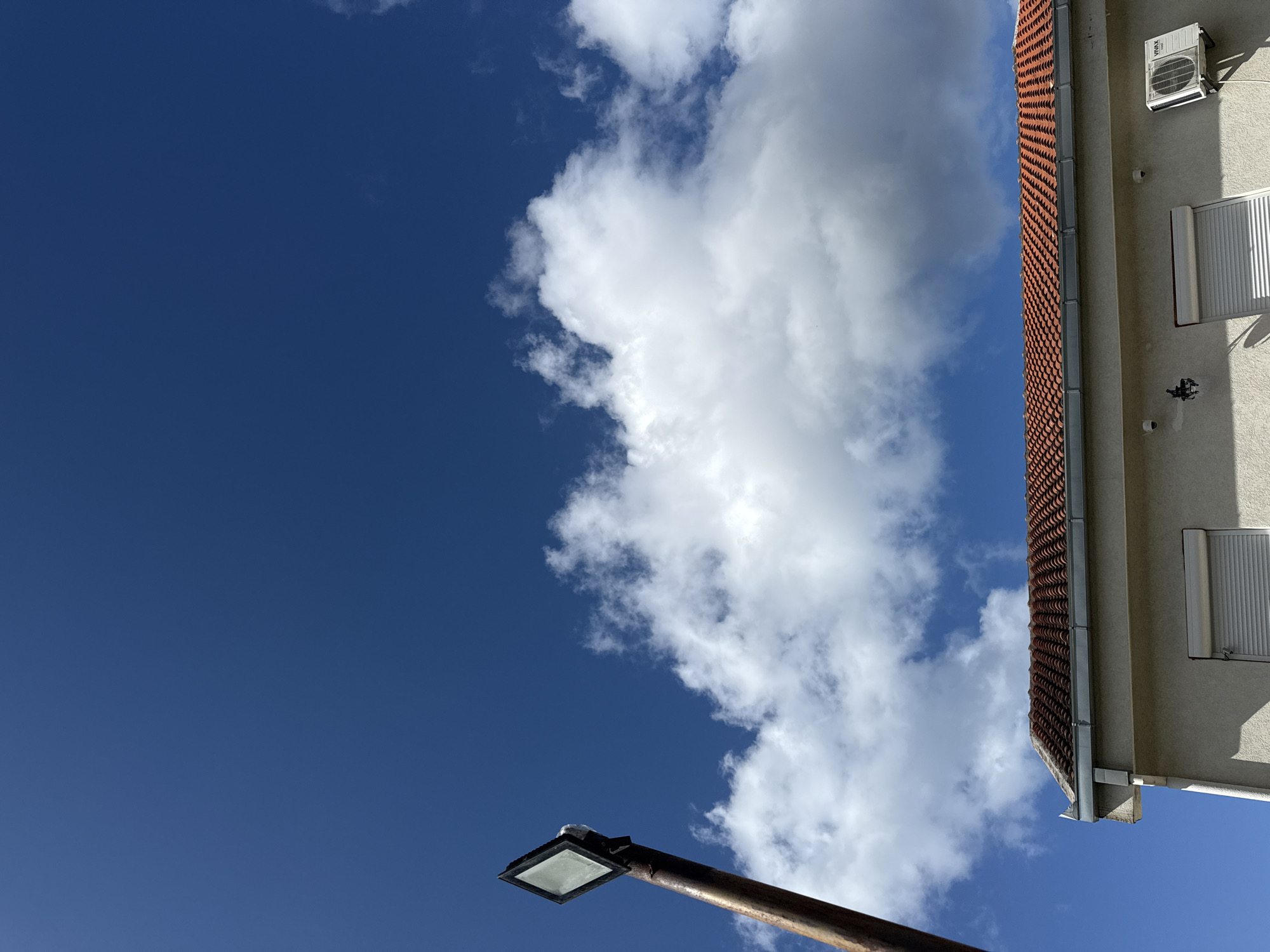 The image captures an outdoor scene dominated by a vibrant blue sky with fluffy white cumulus clouds, set against a partial view of a building and a street light. The bright daylight suggests a clear and sunny day, with distinct shadows visible on the building.
On the right side of the frame, a building with a light-colored, possibly stucco, facade is visible. It features an distinctive orange-red tiled roof with a scalloped pattern, and a grey metal gutter system runs along its edge. High on the wall, a white air conditioning unit is mounted, bearing the brand name "VIVAX." Below it, a white, horizontally slatted roller shutter is closed over a window opening. Several small, round white fixtures are also attached to the building's wall.
The vast expanse of deep blue sky occupies the left and upper portions of the image. A large, bright white cloud mass stretches across the mid-section, appearing dense and well-defined, while smaller, wispy clouds are scattered around.
In the lower-left foreground, a dark, possibly rusty, metal pole of a street light extends upwards. It is topped with a modern, rectangular lamp fixture, featuring a diffused white light panel. The perspective is looking upwards, emphasizing the sky and the architectural elements from below. The location context indicates this scene is in Budva, Montenegro.