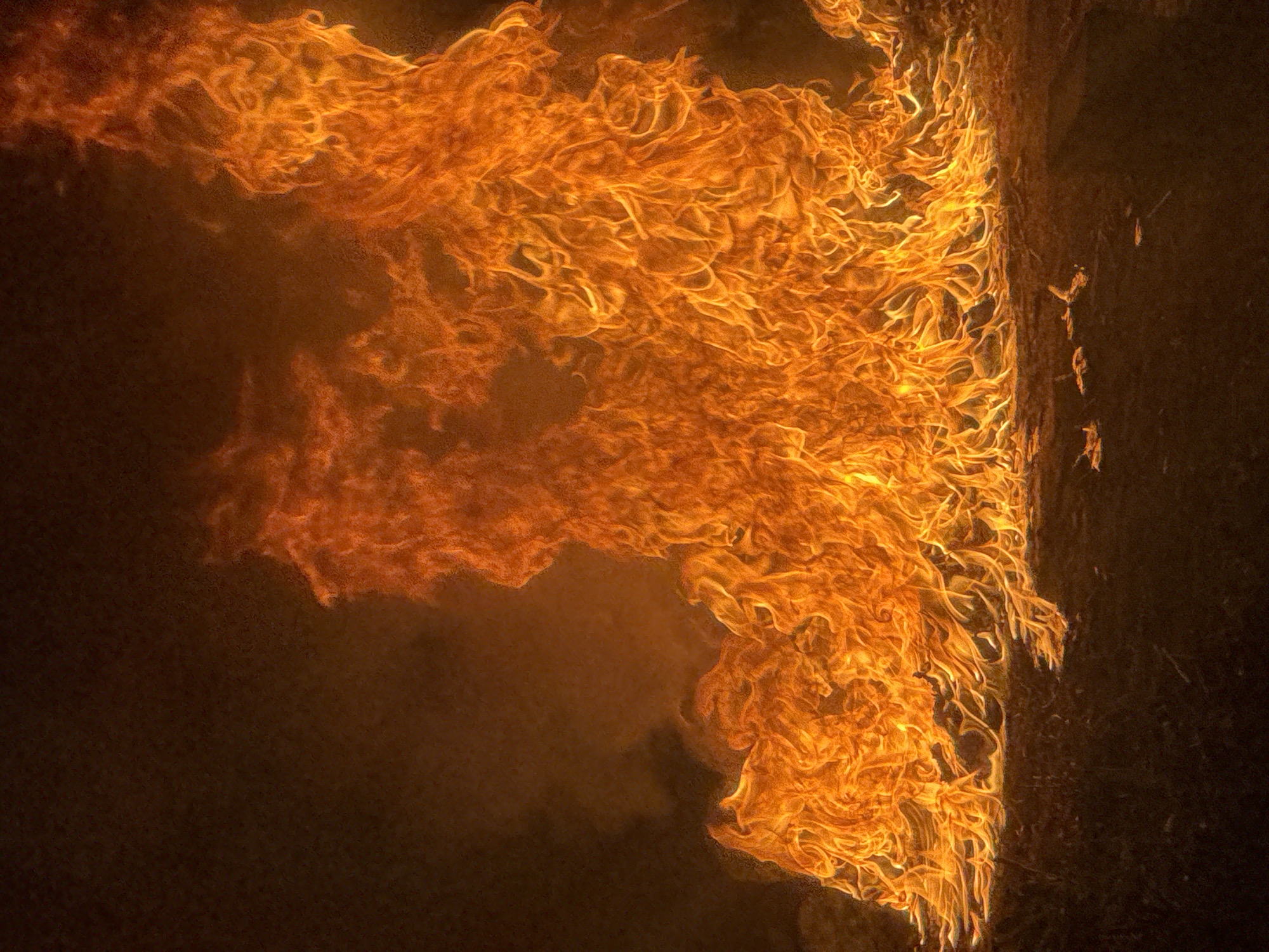 A fire burns intensely on dark ground. Flames, predominantly orange and yellow, rise dynamically from a base that appears to be on the ground. Wisps of smoke are visible in the darker background, creating a hazy atmosphere. The ground itself is dark and appears to be a natural surface, possibly soil or dry vegetation. The overall impression is one of uncontrolled combustion. The location in Жәпек батыр ауылы, Kazakhstan, cannot be determined from the visual information provided in the image.
