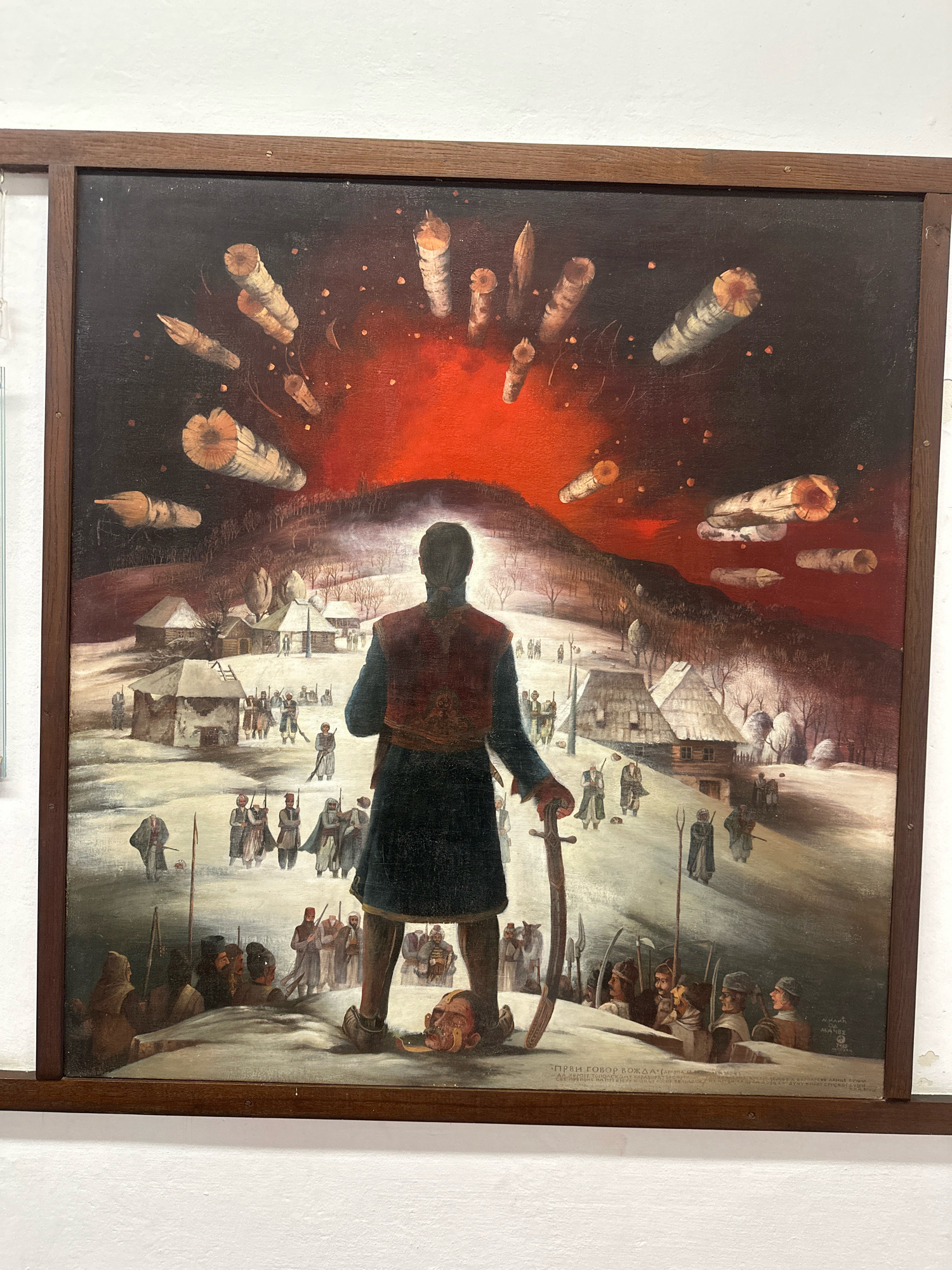 The painting depicts a dramatic scene set in a snow-covered village with a group of figures in the foreground and a fiery, apocalyptic sky in the background. A central figure, seen from the back, stands with a sword, facing away from the viewer and towards the village. This figure is dressed in traditional attire, including a red vest and dark blue coat. Scattered around the village are numerous other figures, some carrying rifles and sabers, suggesting a conflict or gathering. The sky is dominated by an intense red glow, with what appear to be burning logs or projectiles falling from it. The village itself consists of small, traditional houses with thatched roofs, partially covered in snow. The overall atmosphere is one of chaos and intensity. A signature or inscription in Cyrillic script is visible at the bottom right of the painting. The painting is housed in a wooden frame.
The inscription at the bottom of the painting reads: "ПРВИ ГОВОР ВОЖДА" (First speech of the Vožd) and appears to be signed by "П. КАРАНОВИЋ" (P. Karanović). This painting is located at the Oplenac Royal Mausoleum in Topola Municipality, Serbia, specifically in the King Peter's House.