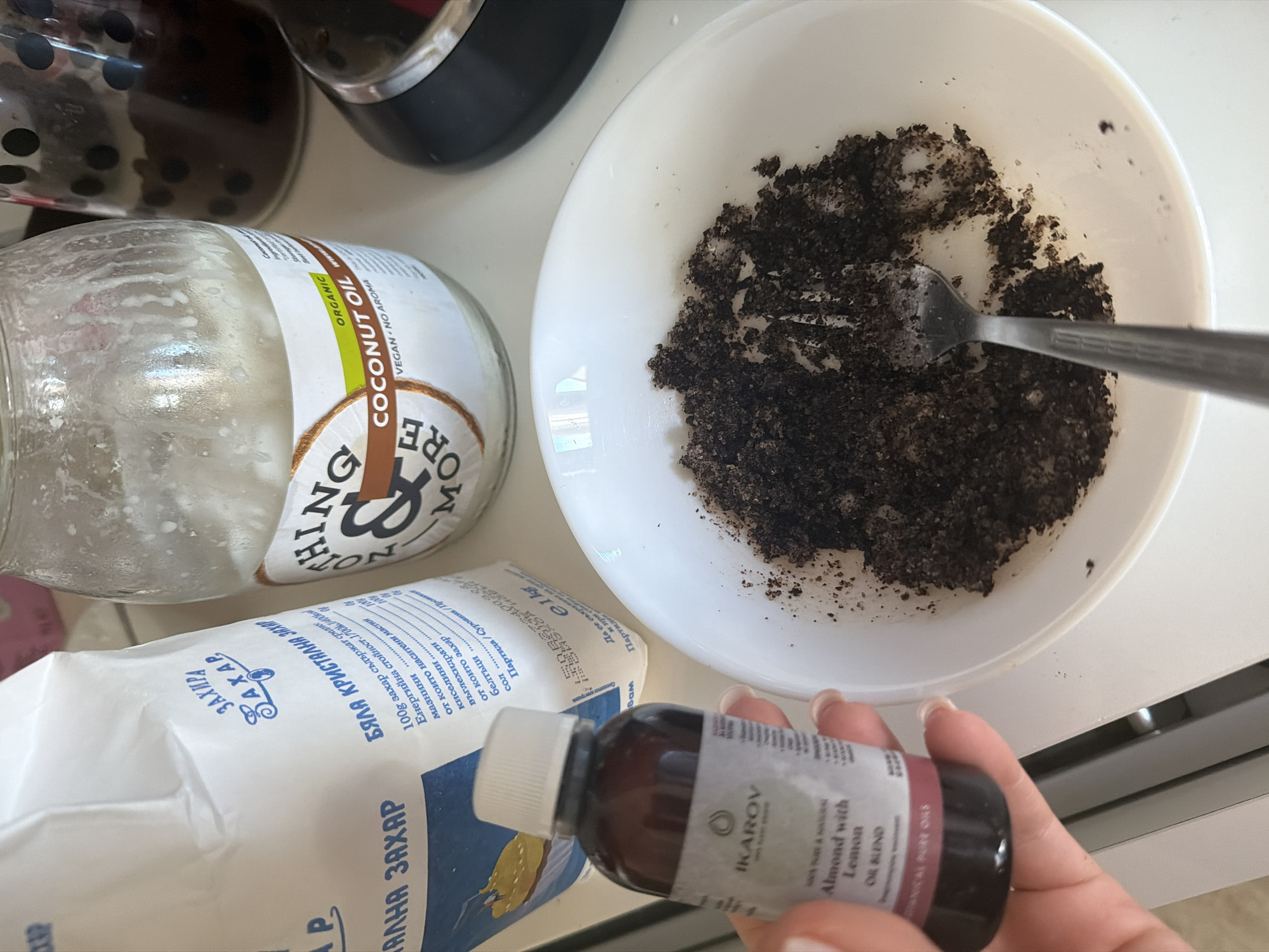 This is a close-up, eye-level shot of a DIY sugar scrub being prepared in a home kitchen. The setting appears to be an indoor countertop, likely in an apartment or house. While the specific location in Sofia, Bulgaria cannot be determined from the image alone, the presence of Bulgarian text on the sugar bag ("БЯЛА КРИСТАЛНА ЗАХАР") strongly suggests this.
In the foreground, a person's hand, with neatly manicured nails, holds a small dark glass bottle labeled "IKAROV Almond with Lemon OIL BLEND". To the right of the bottle, a white ceramic bowl contains a dark, granular mixture, presumably the sugar scrub. A silver fork is partially submerged in the mixture, indicating an active mixing process.
In the background, a large white paper bag with blue lettering, also in Bulgarian ("ЗАХАР"), is visible. Next to it stands a glass jar containing a white, semi-solid substance, labeled "ORGANIC COCONUT OIL VEGAN - NO AROMA". A partially obscured, dark glass object with black dots is visible behind the coconut oil jar. The overall lighting is bright, suggesting natural light from a window, and the surfaces appear clean and organized.