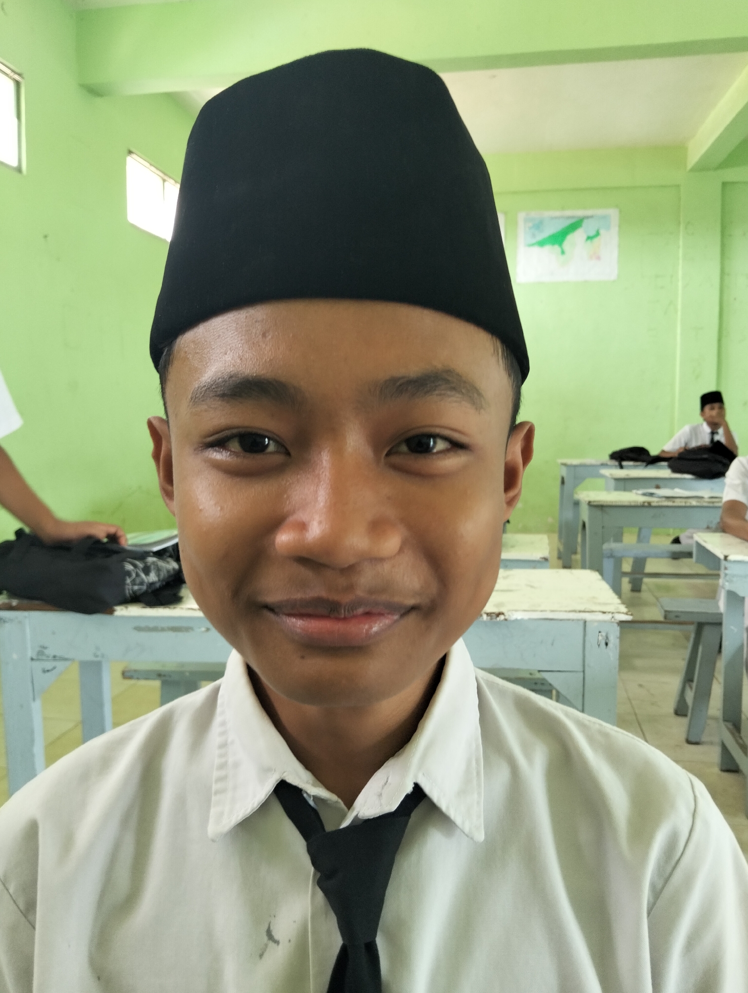 A young Indonesian male with dark skin and short black hair is pictured from the chest up, wearing a white collared shirt, a black tie, and a black kufi. He is looking directly at the camera with a slight smile. The background shows a classroom with green walls, white ceiling, and rows of light blue desks. In the distance, another young male student wearing similar attire is visible sitting at a desk. A hand is seen reaching for a black bag on a desk in the foreground to the left. The setting appears to be a school in Kranji, Indonesia, based on the context of the image's potential origin.