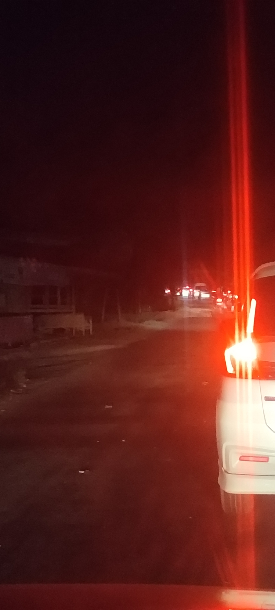 A nighttime scene on a road in Meuse, Indonesia. In the foreground, the rear of a white car is visible, with illuminated red taillights. The road stretches into the distance, and in the background, there are other vehicles with their lights on, suggesting traffic. Buildings are barely discernible on the left side of the road. The overall lighting is dim, predominantly red due to the car lights and ambient conditions.