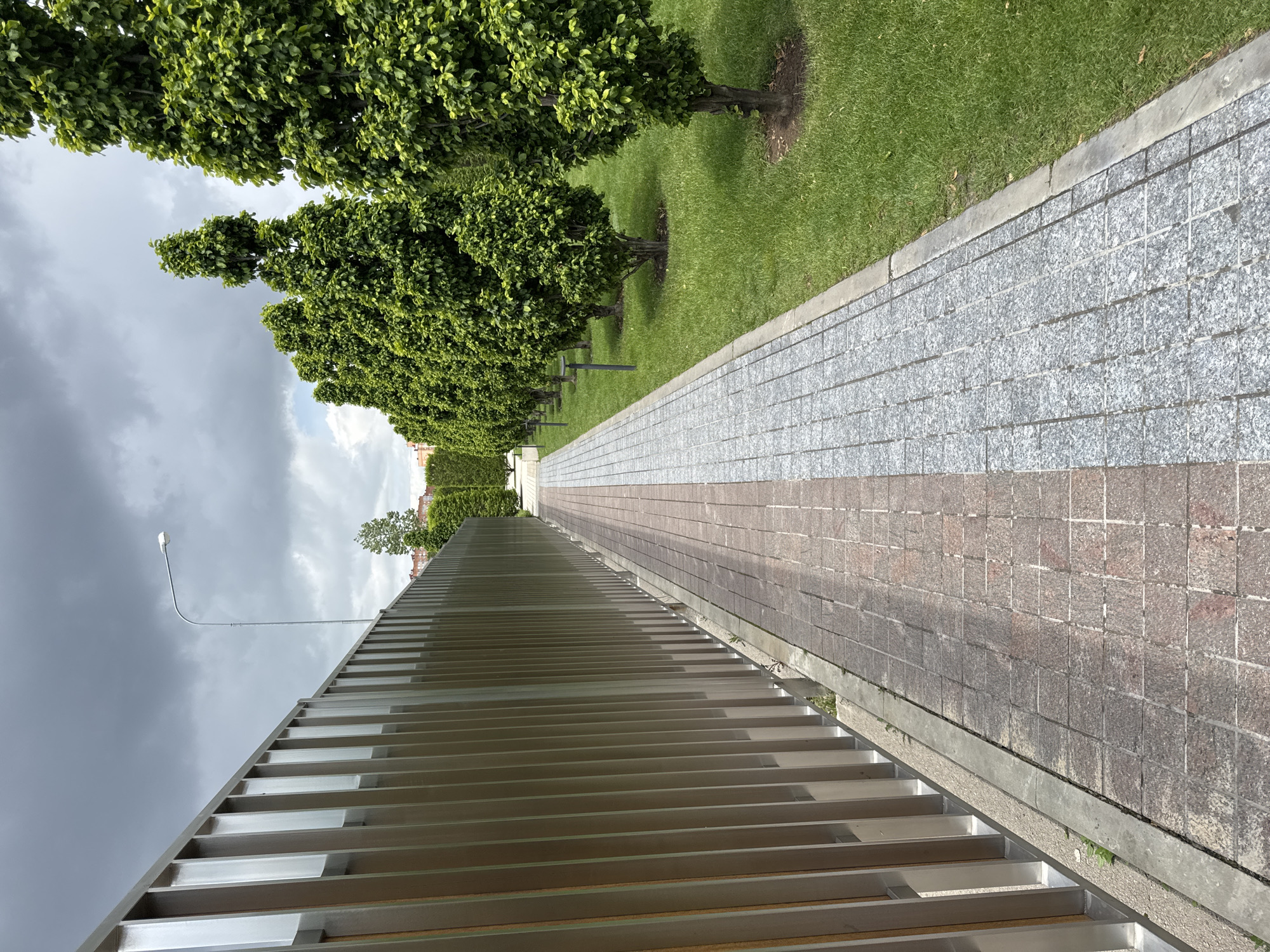 The image depicts a walkway in what appears to be a public park or pedestrian zone. The walkway is constructed of two types of paving stones: a gray granite-like material and a red brick-like material laid in a rectangular pattern. Adjacent to the walkway is a strip of green grass with evenly spaced, pruned trees planted along its edge. On the opposite side of the walkway is a metal barrier or fence consisting of horizontal bars. The sky overhead is overcast with dark storm clouds gathering. A lone streetlight is visible extending upward from behind the metal structure. The location is Krasnodar, Russia. No people or subjects are visible in the image. There are no observable actions or interactions taking place.
