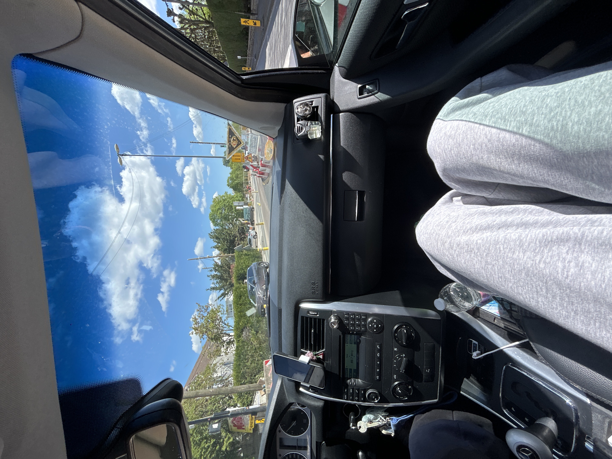 The image, taken from inside a car, shows a view of a street in Noisy-le-Grand, France. The car is stopped at what appears to be a road construction zone, indicated by orange and white barriers and warning signs. Visible through the windshield is a street scene with buildings, trees, and other vehicles. The sky is blue with scattered clouds. Inside the car, the dashboard and parts of the driver's seat, including a person wearing gray sweatpants, are visible. A mobile phone is mounted on the dashboard. A set of keys and a water bottle are visible in the center console.