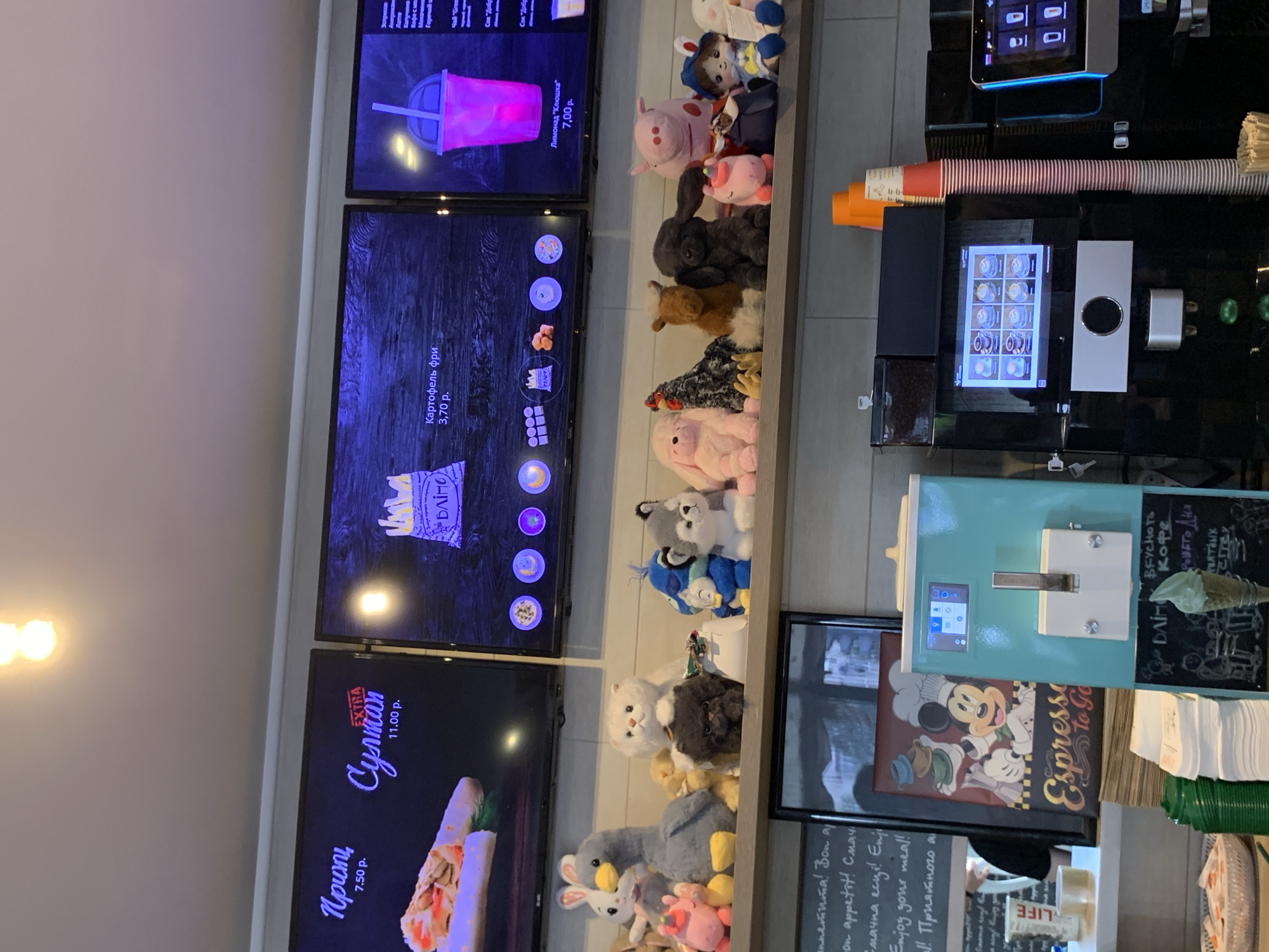 The image shows a close-up of a counter display at a food establishment, likely a cafe or fast-food restaurant, with a focus on plush toys and menu screens. In the background, two large screens display menu items. The left screen shows a shawarma-like wrap labeled "Гриппы 7.50 р." and a dish labeled "Супман EXTRA 11.00 р.". The right screen displays a pink beverage in a cup labeled "Лимонад \"Клоушка\" 7.00 р.". Below the screens, a shelf is lined with an assortment of plush toys, including a pink pig, a goose, a white cat, a fluffy black cat, a dog, a unicorn, a chicken, a brown animal resembling a cow, a duck, a husky, a doll with a blue hat, and other various animals.
In the foreground, a turquoise soft-serve ice cream machine is visible, with a Mickey Mouse themed poster to its left. To the right of the ice cream machine, a commercial coffee machine with a touchscreen display is present, and stacks of paper cups are arranged next to it. A single ice cream cone with green ice cream sits on the counter in front of the coffee machine.
The overall setting appears to be indoors, with a tiled wall in the background. The establishment is likely located in Sluck, Belarus, as indicated by the menu items being in Cyrillic script and common offerings at such establishments. No people are clearly visible in the frame. The lighting suggests an indoor environment, possibly with overhead lighting.