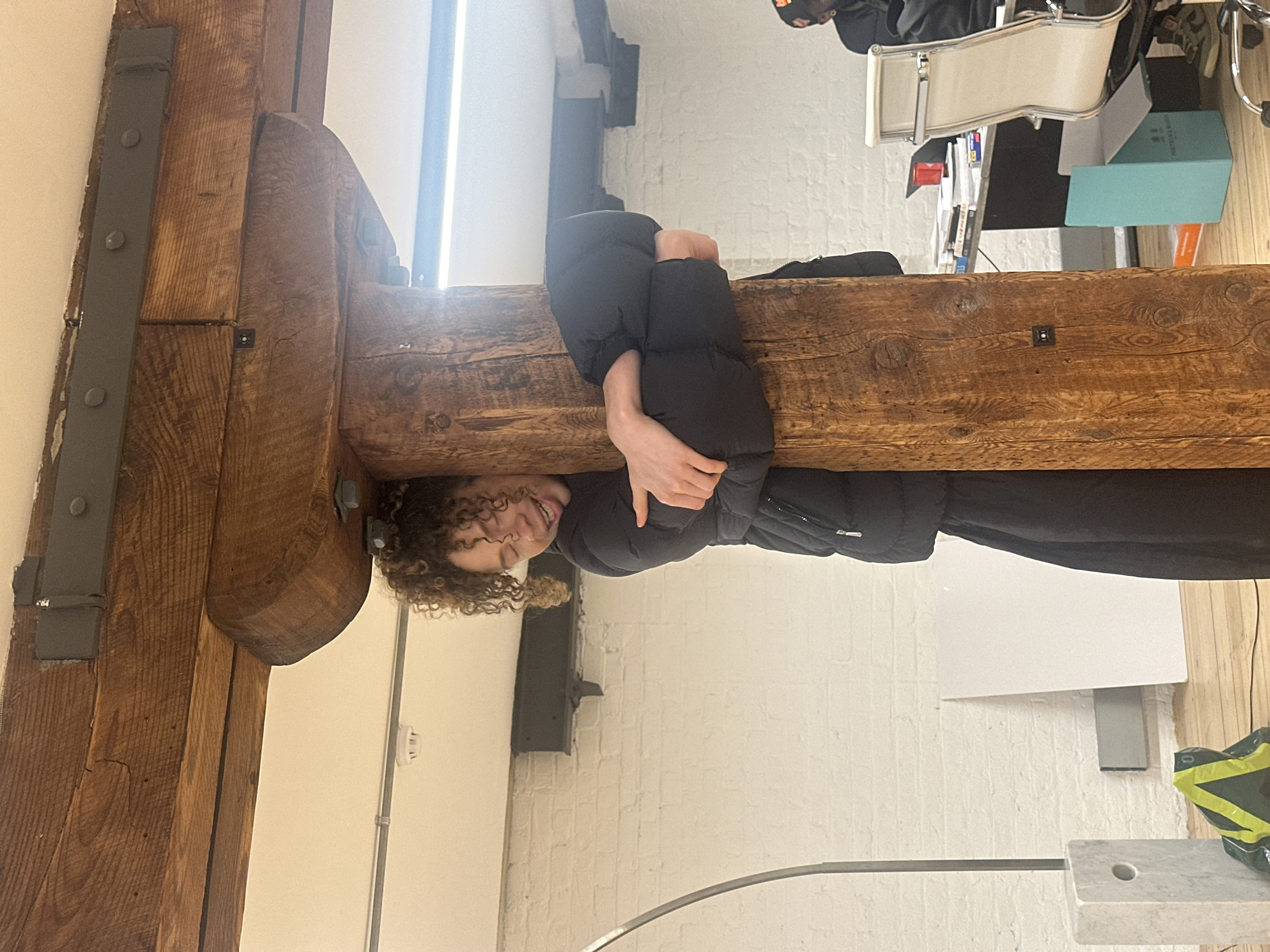 The image displays an indoor scene featuring a young woman playfully positioned on a large, horizontal wooden beam. The woman, with light brown curly hair, is wearing a black puffer jacket and black pants. She is seen from the chest up, with her arms wrapped around the beam, and her head turned towards the viewer, revealing a wide, joyful smile or laugh.
The wooden beam, a prominent feature in the foreground, is robust and appears aged, showcasing distinct wood grain patterns and knots. It is structurally reinforced with dark grey or black metal plates secured by visible rivets or bolts, particularly noticeable on the left side of the image where the beam meets a wall.
In the background, a textured white brick wall is visible, particularly below the beam and extending to the right. To the far left, a bright vertical strip suggests a window or a tall light fixture, creating a bright glare in that area. Above the beam, towards the right, an office-like setting is discernible, including a desk, a white office chair with a metal frame, and several items on the desk such as books or files, one of which is a light blue color. A green and yellow bag can be seen on the floor to the right. Further right, a modern-looking floor lamp with a curved metal arm and a white marble base is partially visible. A white smoke detector or similar utility device is mounted on the white wall in the upper left background.
The overall atmosphere of the image is lighthearted and playful, primarily conveyed by the woman's smiling expression. The lighting appears bright and natural, indicating daytime. No discernible text is visible on any objects or surfaces.