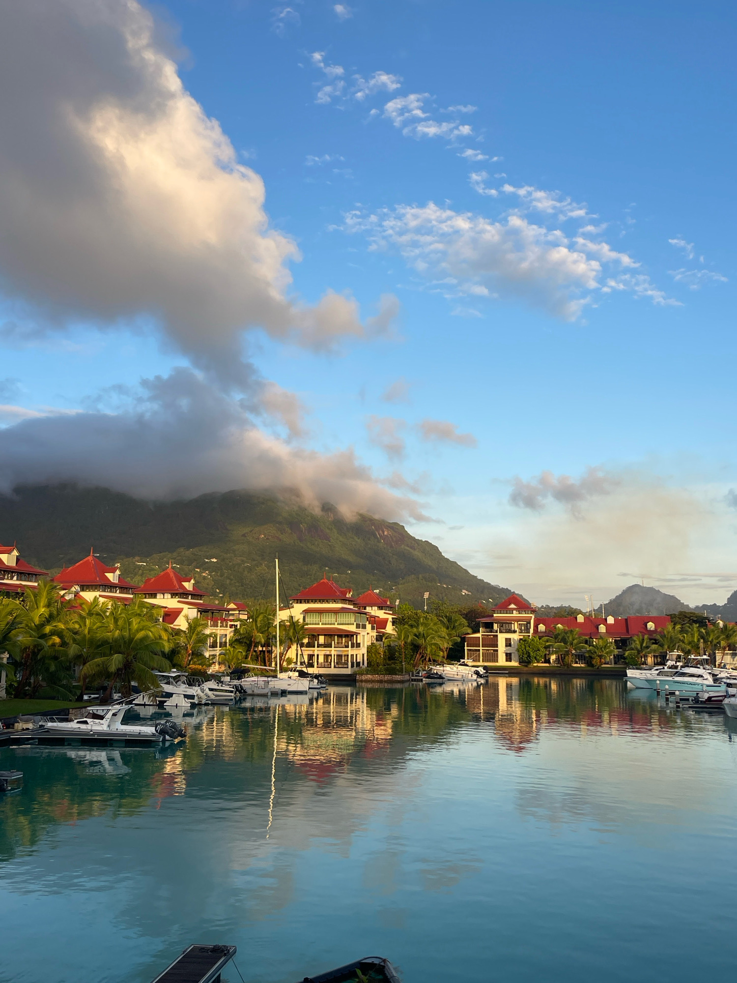 The image depicts a scenic harbor scene in Providence, Seychelles. In the foreground, calm, turquoise water reflects the sky and the surrounding architecture. Several white motorboats are docked in the marina, their hulls casting reflections on the water. Behind the boats, a row of buildings with distinctive red roofs and cream-colored facades lines the waterfront. These structures are set against a backdrop of lush green palm trees and a verdant, mist-shrouded mountain range. The sky above is a vibrant blue, dotted with fluffy white clouds and some larger, more ominous clouds lingering over the mountains, hinting at the possibility of changing weather. The overall impression is one of tropical tranquility and resort living.