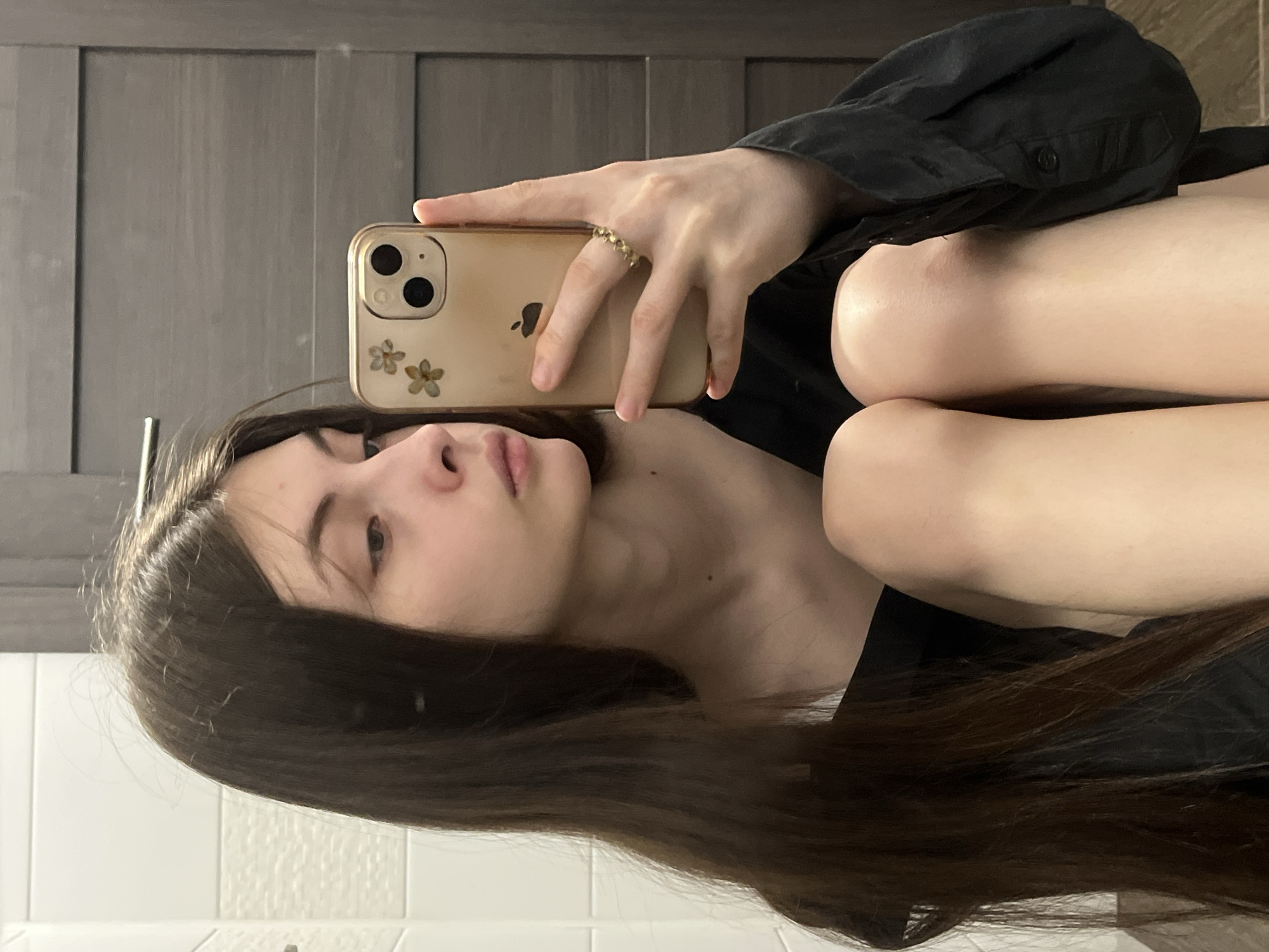 A young woman is taking a selfie in what appears to be a bathroom. She is holding an iPhone in her right hand, which is adorned with a ring. The phone case is beige with flower decorations. She is wearing a dark shirt. Her long brown hair falls across her shoulder. The background includes a gray paneled door and white tiled walls. There is no evidence to suggest the location is Saint Petersburg, Russia.
