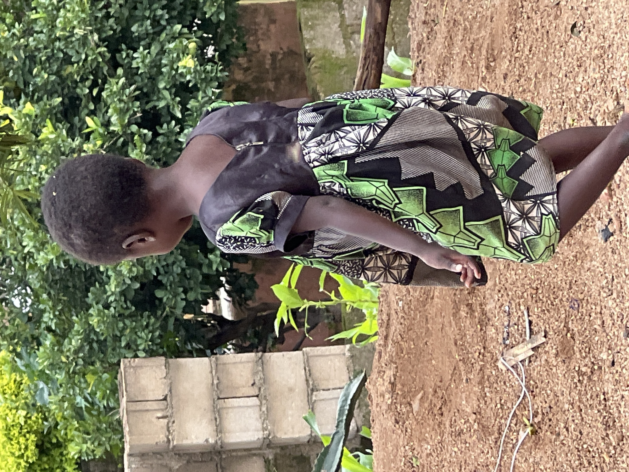 In a dusty, outdoor environment that appears to be within the city of Jos, Nigeria, a young child stands facing away from the camera. The child is wearing a dark, possibly black, top with a zipper closure and a short-sleeved dress with a vibrant, geometric pattern featuring green, black, and white. The dress has a distinct African print. The background consists of lush green foliage and a partially constructed brick wall. The ground is covered in reddish-brown dirt and small stones. A thin, white wire is visible on the ground near some wooden debris. The scene suggests a casual, everyday setting.